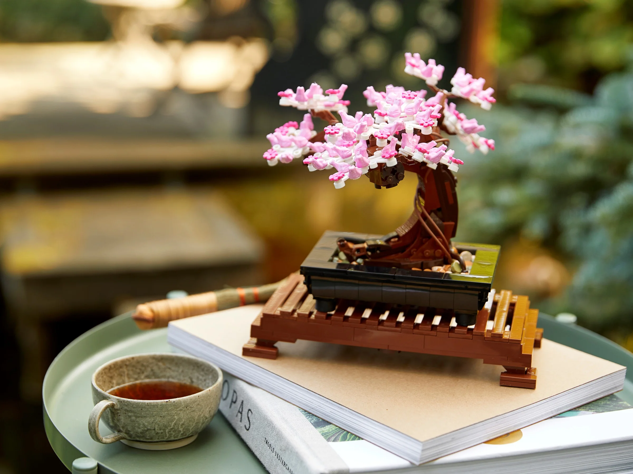 Bonsai LEGO aranjat artistic cu flori roz pe o măsuță, lângă o ceașcă cu ceai.