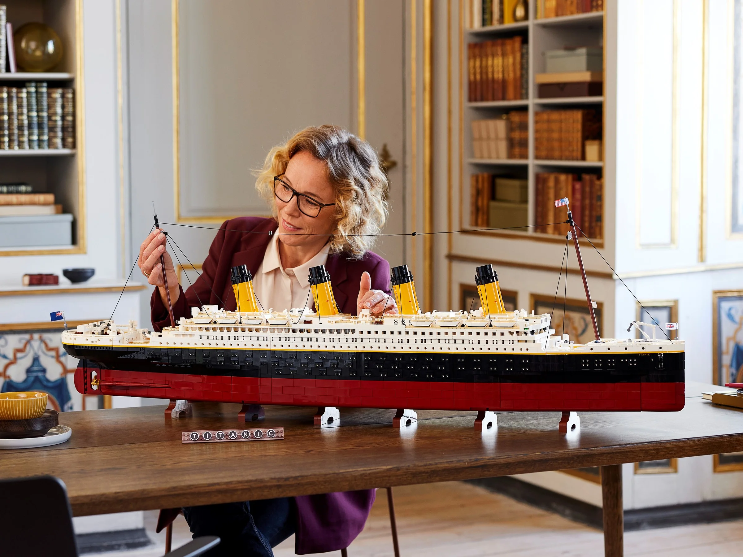 Femeie asamblează modelul LEGO Titanic, afișat pe o masă într-o cameră elegantă. Complexitate și detaliu.