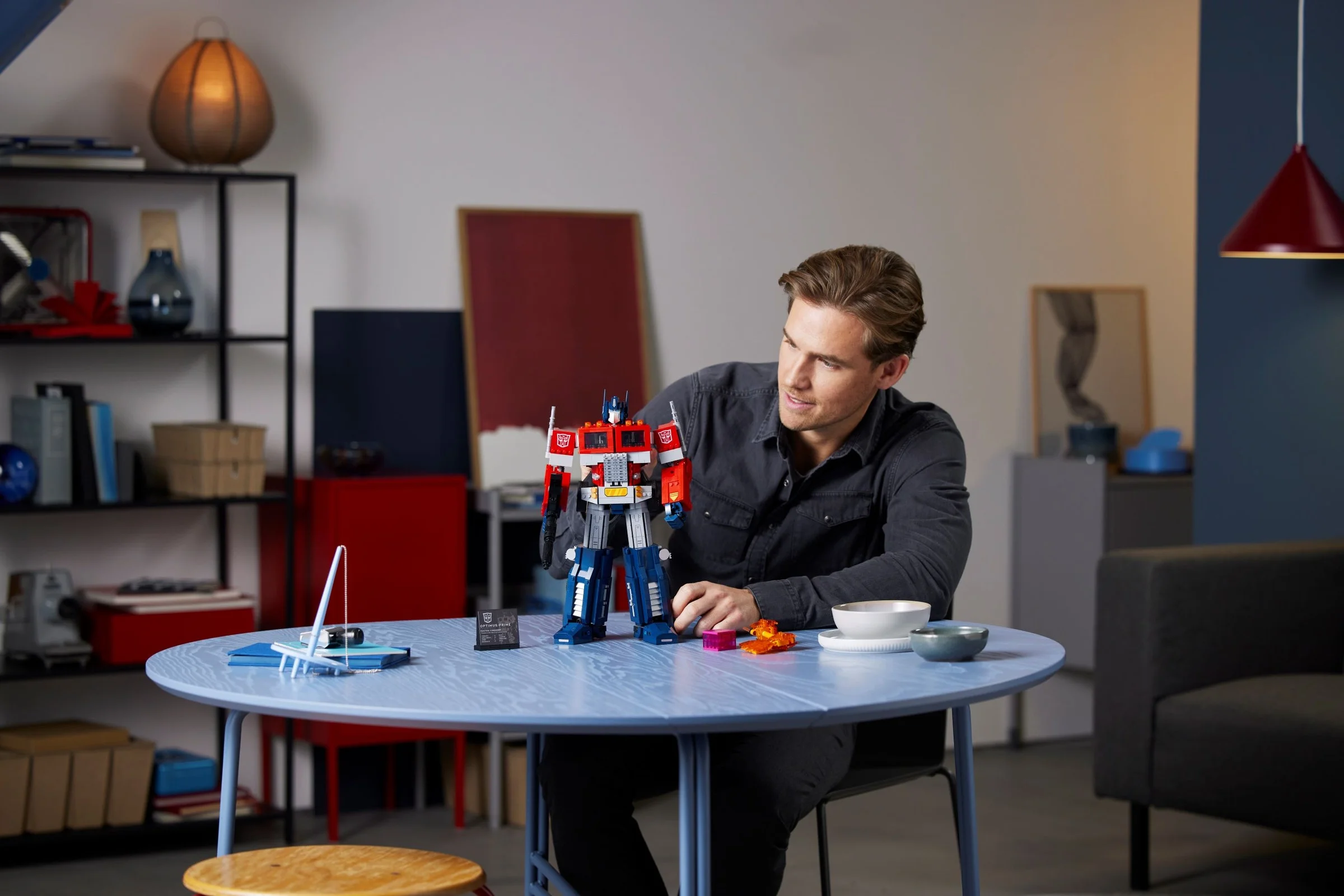 Un bărbat admiră Optimus Prime LEGO pe o masă albastră, într-un cadru interior modern.
