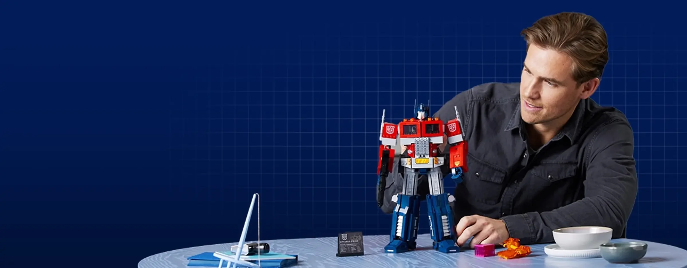 Modelul LEGO Optimus Prime alături de un adult, pe o masă, ilustrând interacțiunea cu produsul.