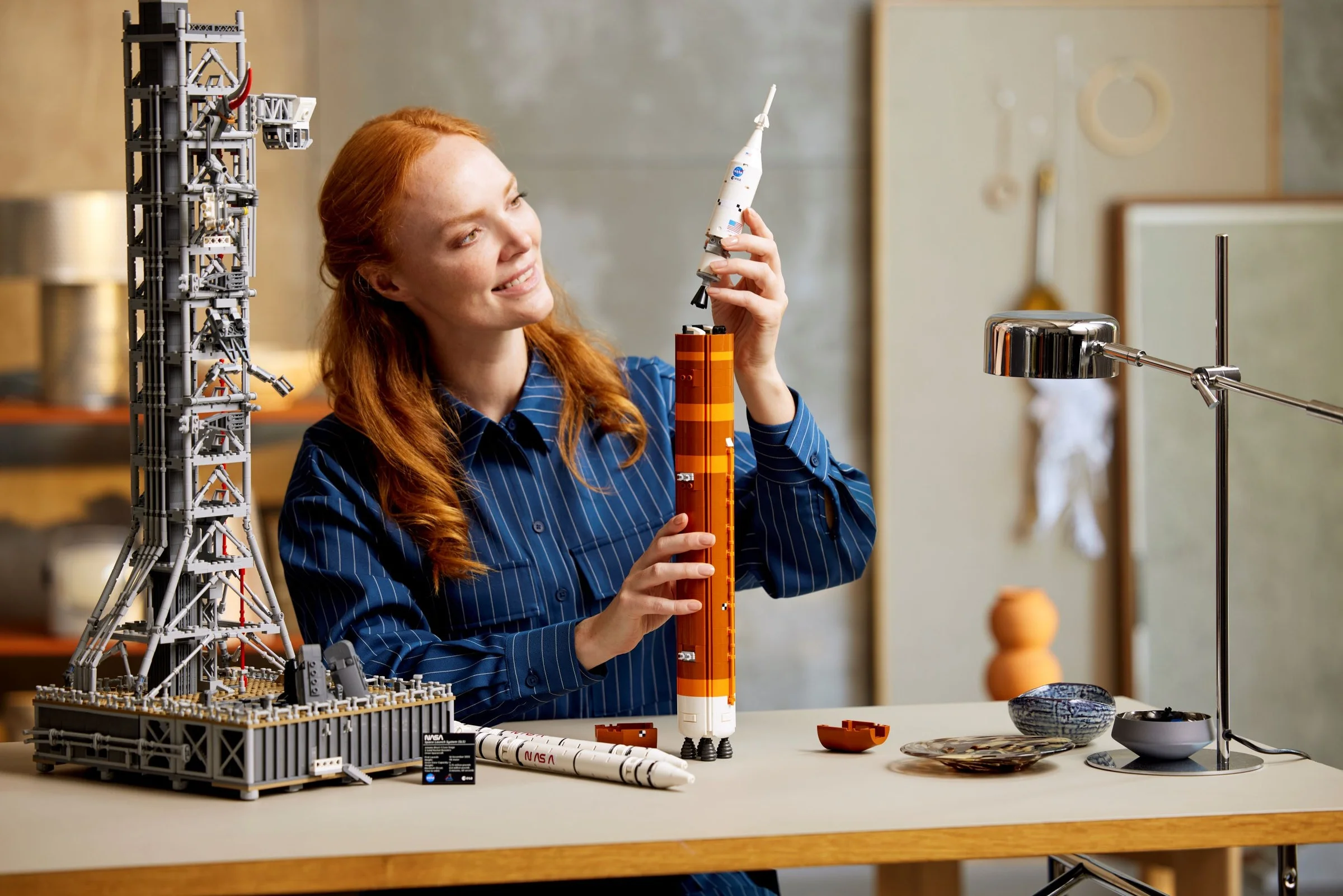 O femeie asamblează racheta Artemis LEGO, zâmbind. Construcția include turnul de lansare, racheta și propulsoarele.