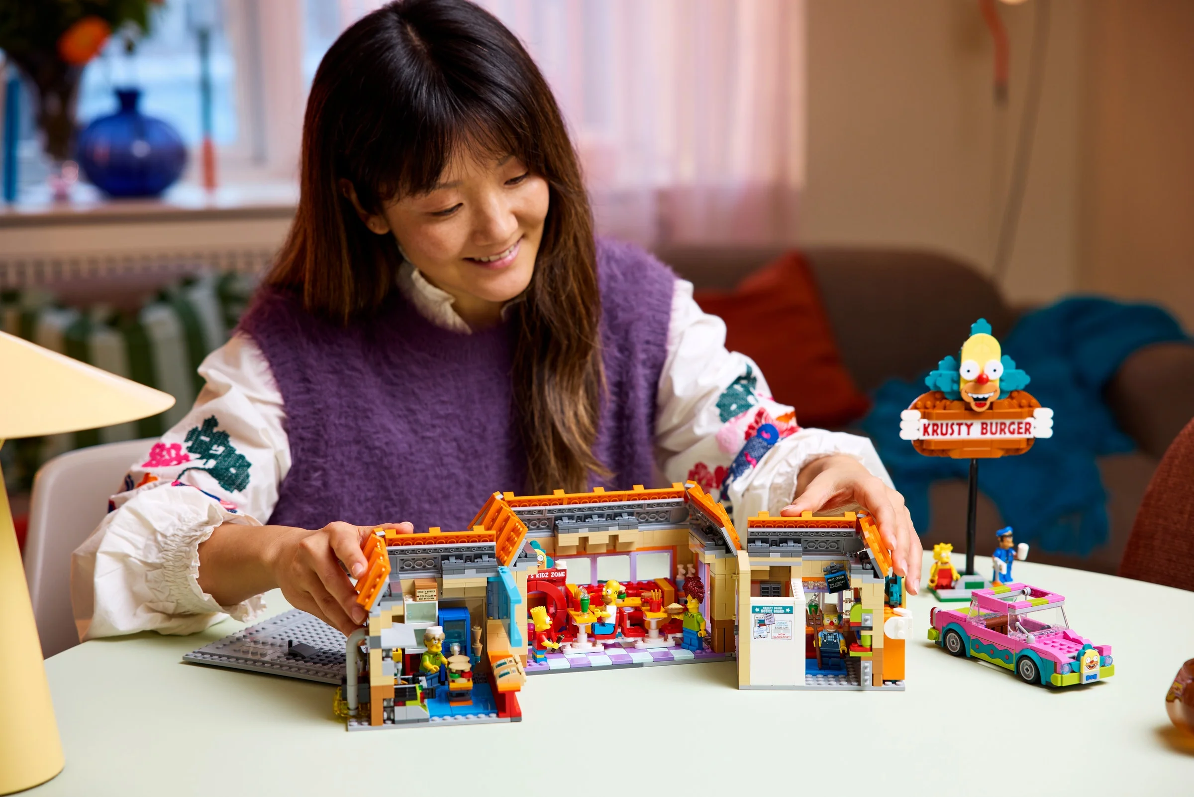 Femeie construind Krusty Burger LEGO. Set detaliat, minifigurine și mașină vizibile.