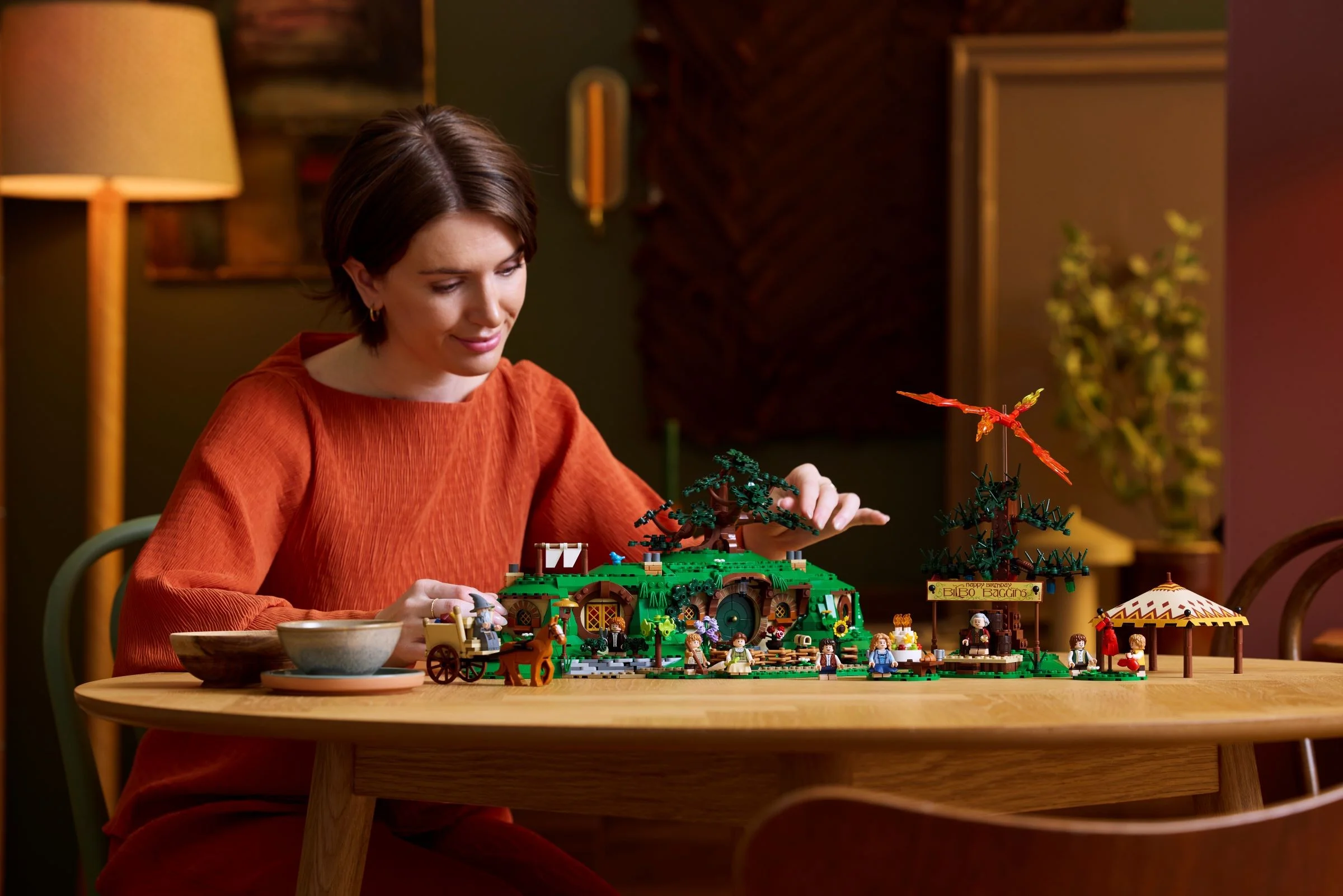 O femeie se joacă cu setul LEGO Hobbiton pe o masă din lemn. Atmosferă primitoare și detaliată.