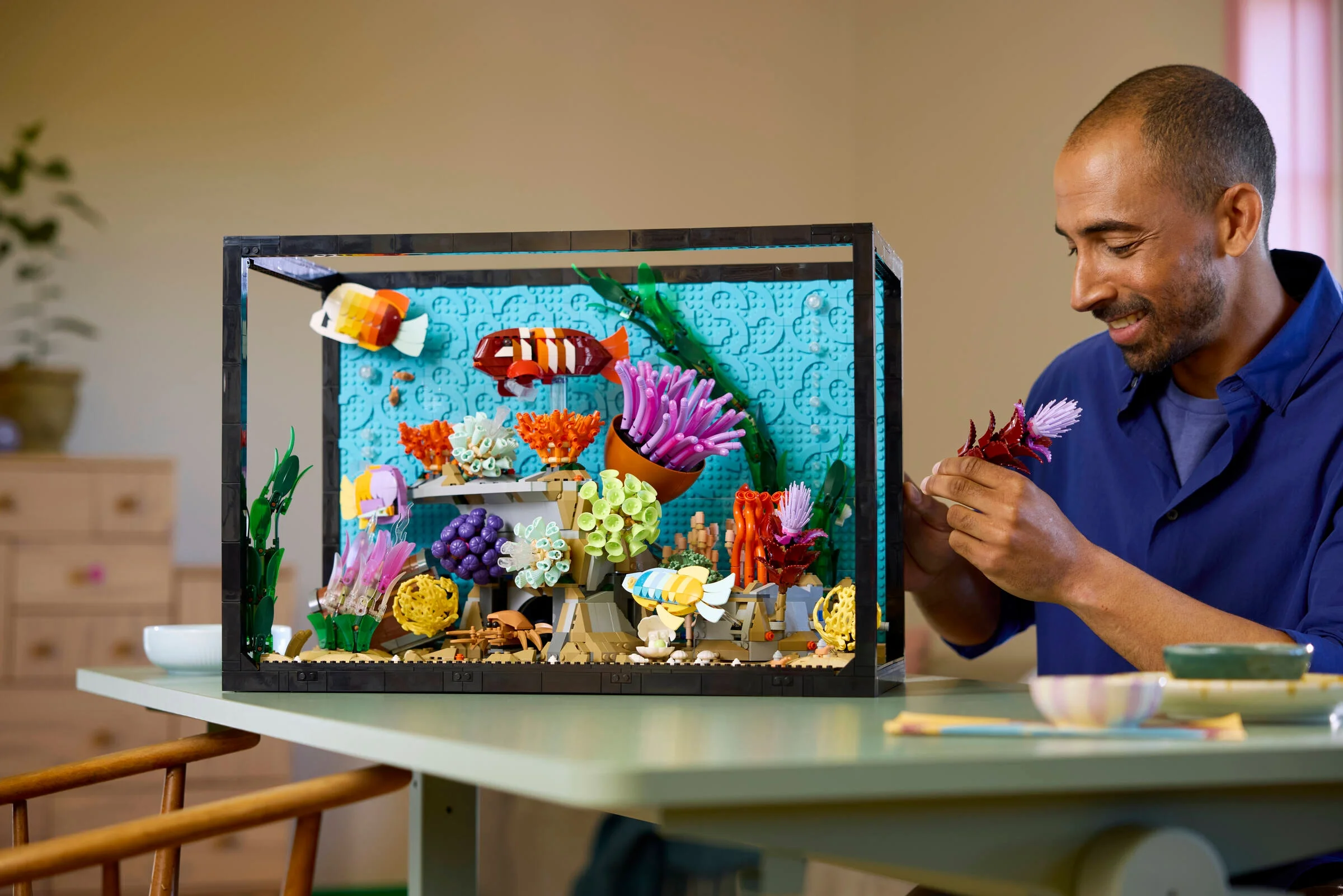Un bărbat construiește un acvariu tropical LEGO cu pești și corali colorați.