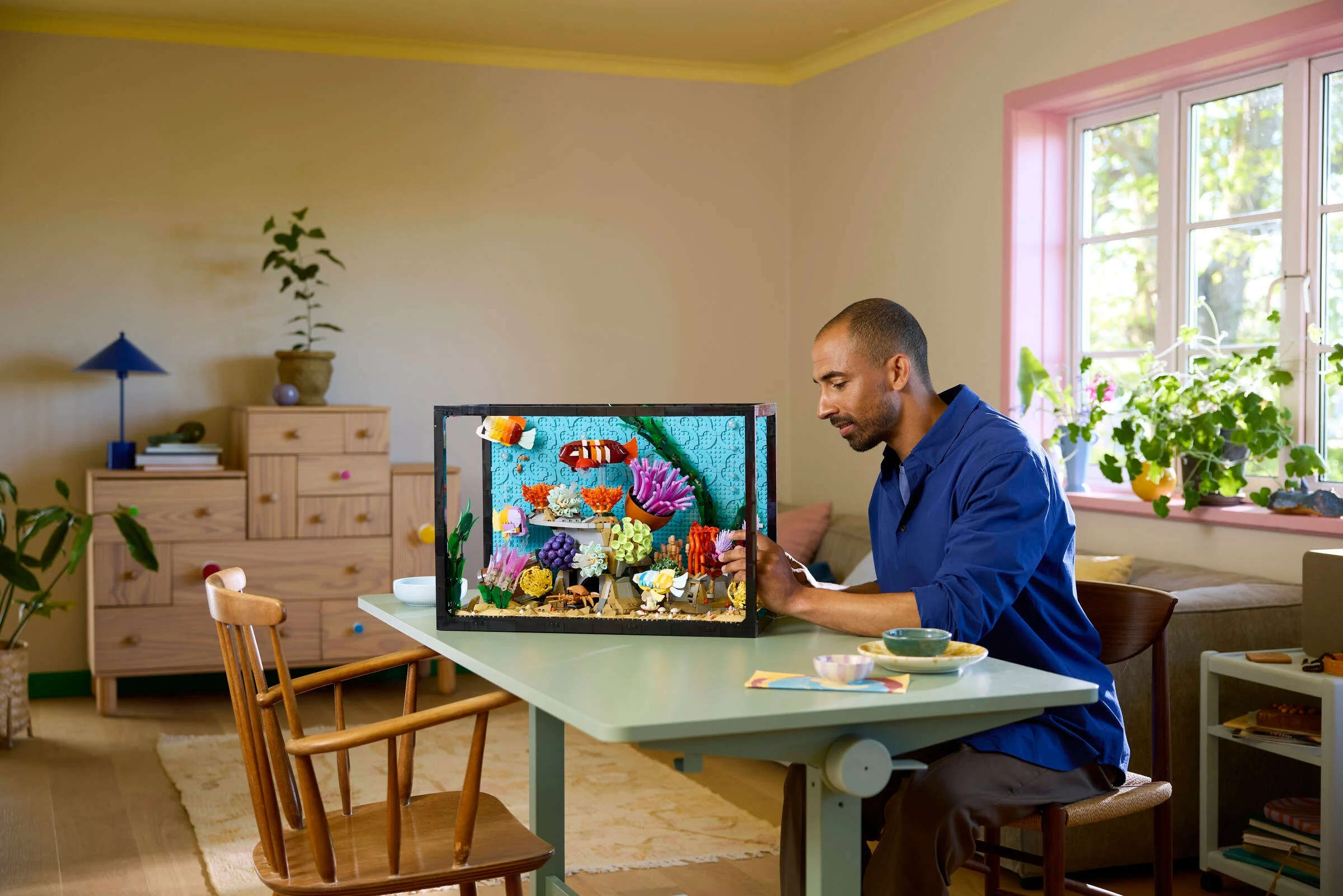 Un bărbat construiește acvariul LEGO 10366 într-o cameră luminoasă, cu mobilier colorat.