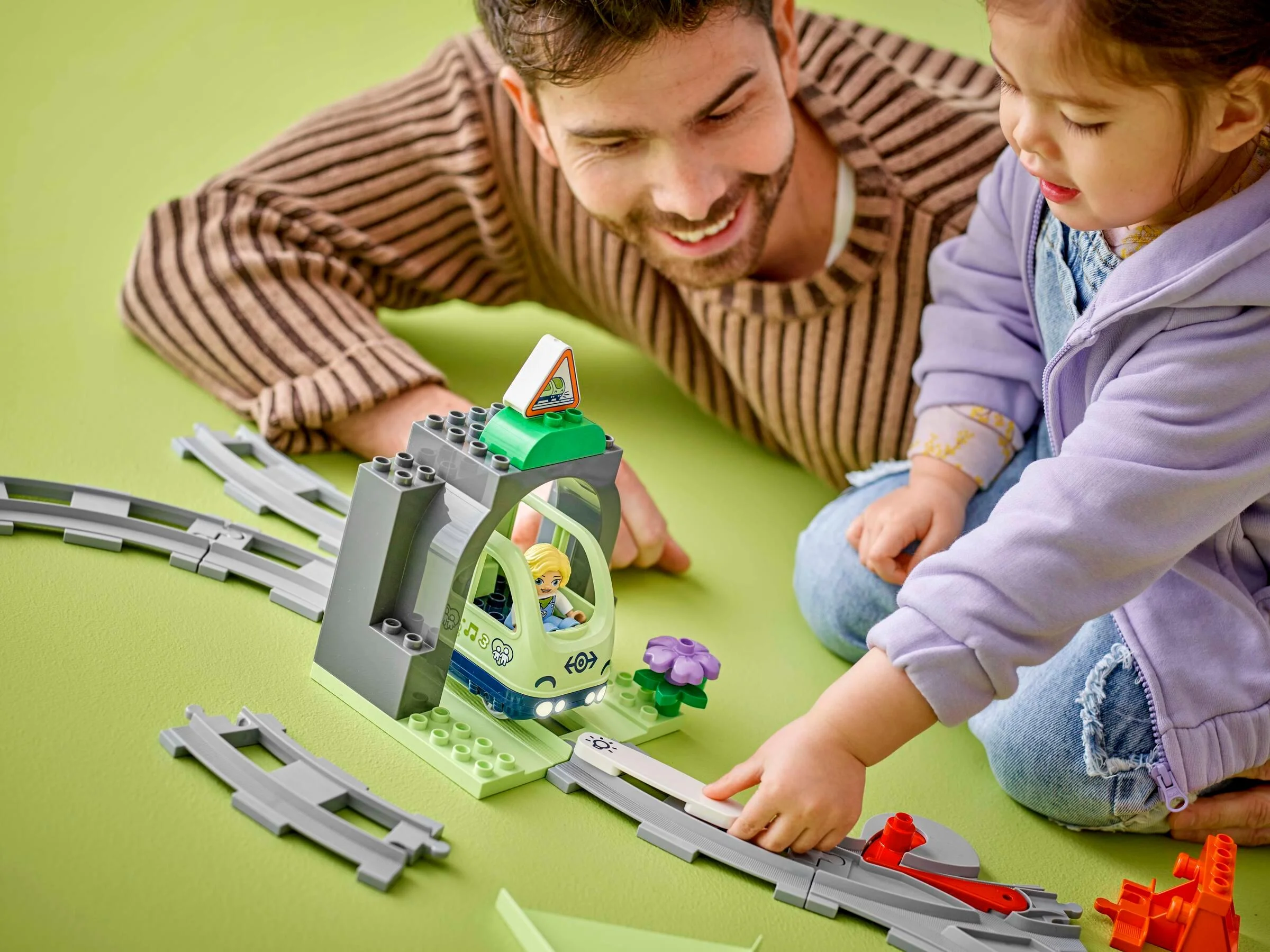 Un tată zâmbitor și fiica sa se joacă cu trenul Duplo și tunelul pe șine, într-o scenă caldă și educativă.
