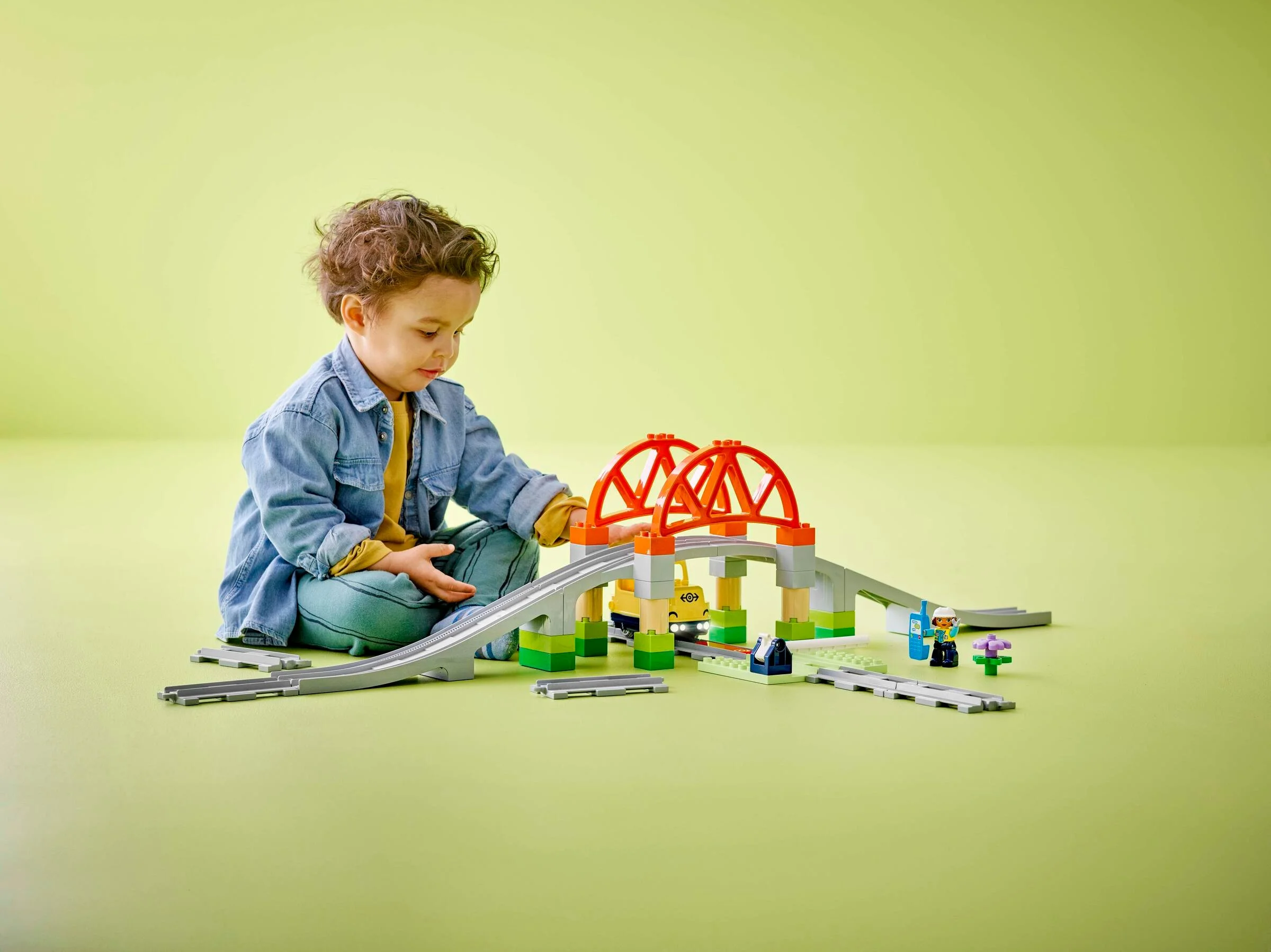 Copil se joacă cu trenul LEGO Duplo, cu pod, șine și minifigurine.