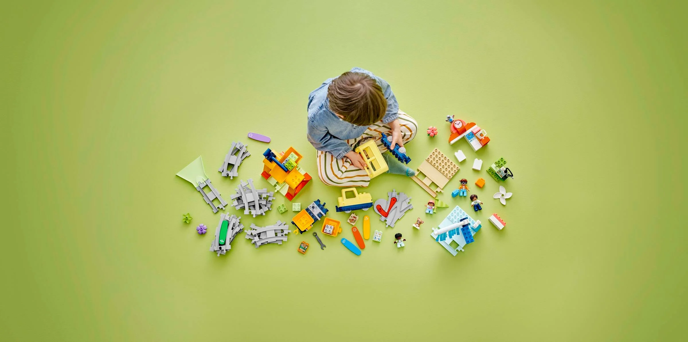 Copil se joacă cu setul LEGO Duplo Trenul Comunității. Piese împrăștiate pe verde.