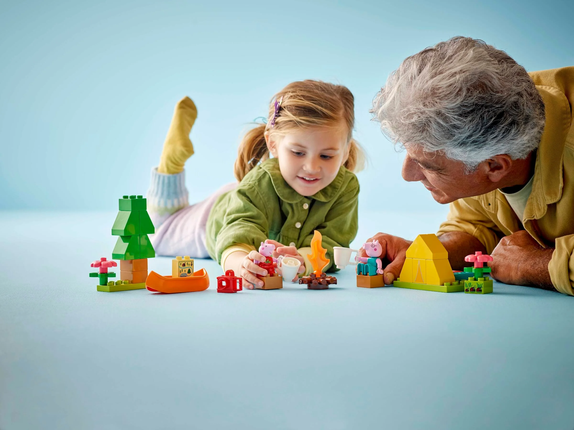Fetiță și adult se joacă cu setul LEGO Duplo Camping Trip, creând o scenă cu cort și foc de tabără.