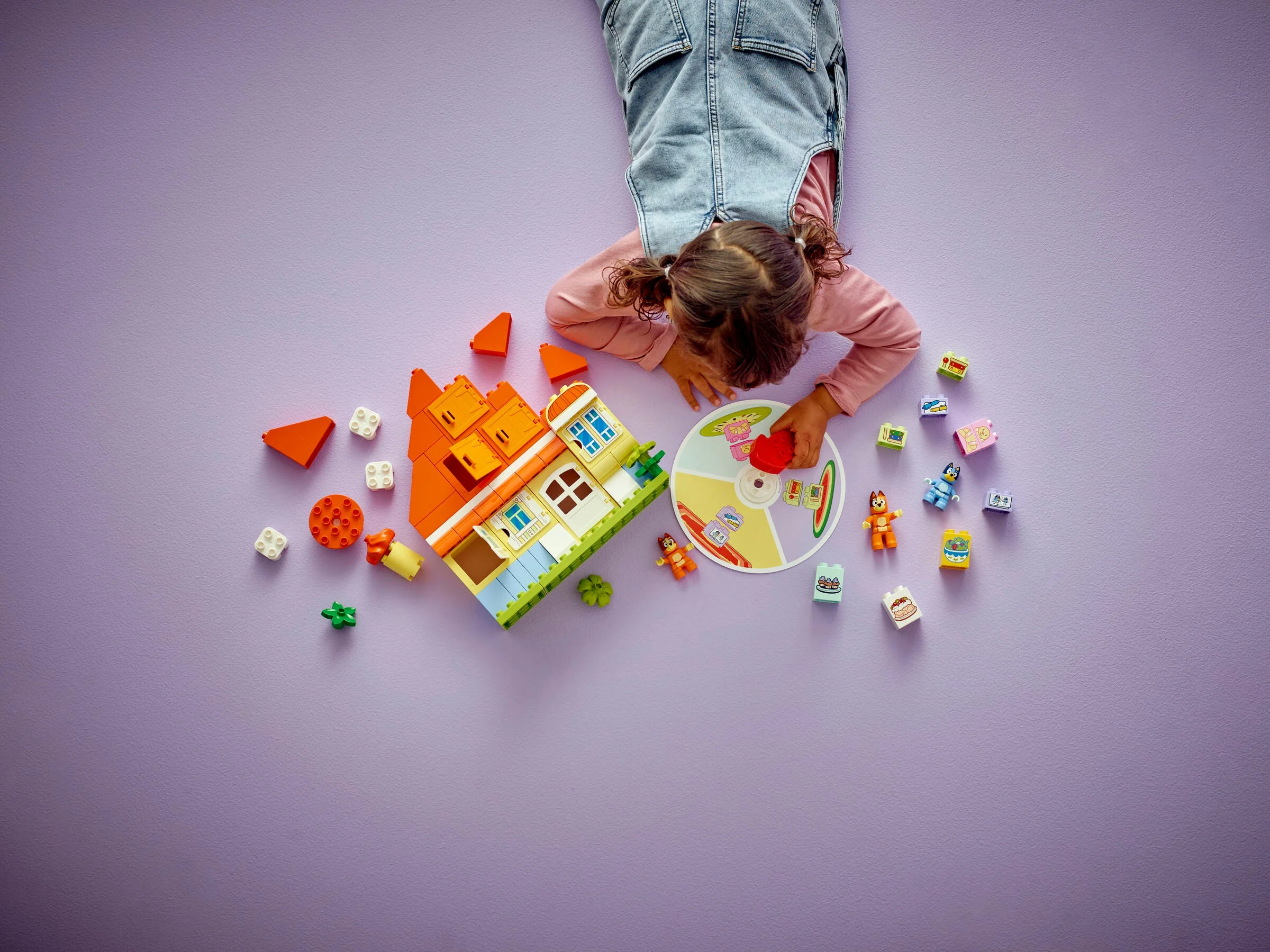 O fetiță se joacă cu setul LEGO Duplo Bluey's Family House și jocul de memorie pe o suprafață violetă.