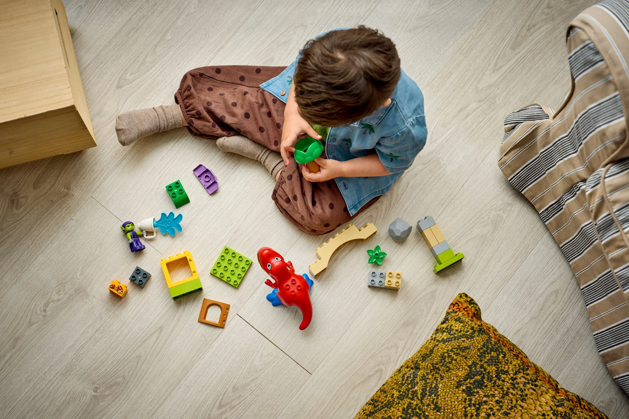 Copil se joacă cu setul LEGO Duplo Spidey-Rex vs. Green Goblin pe podea. Blocurile sunt colorate și variate.