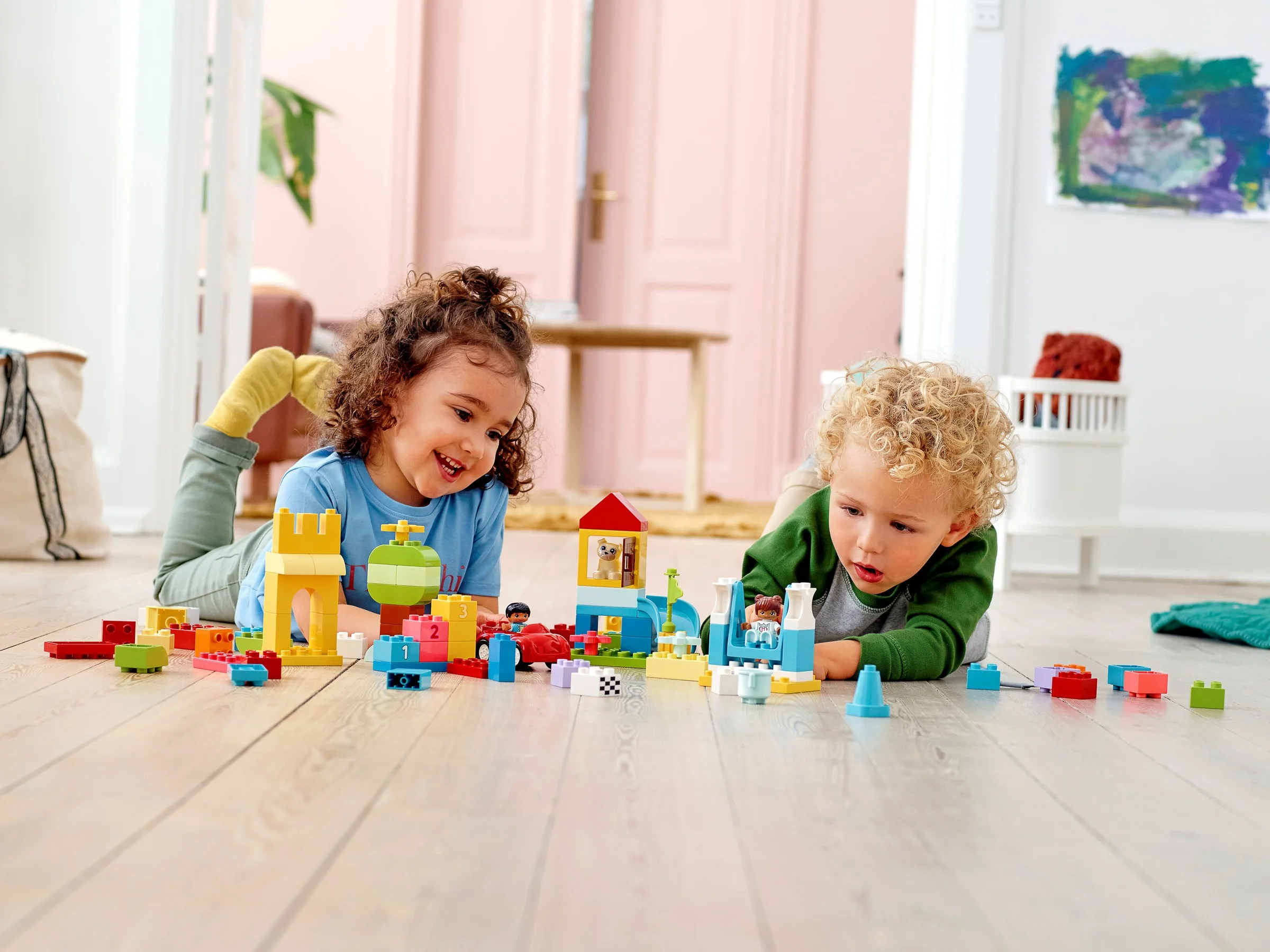 Doi copii se joacă cu cărămizi Duplo colorate, zâmbind și construind structuri creative.
