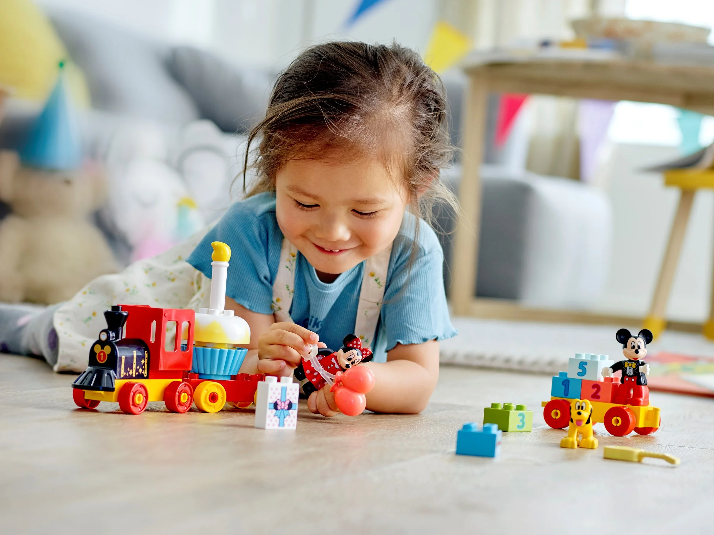 O fetiță zâmbește jucându-se cu trenulețul aniversar Mickey și Minnie Duplo, pe podea.