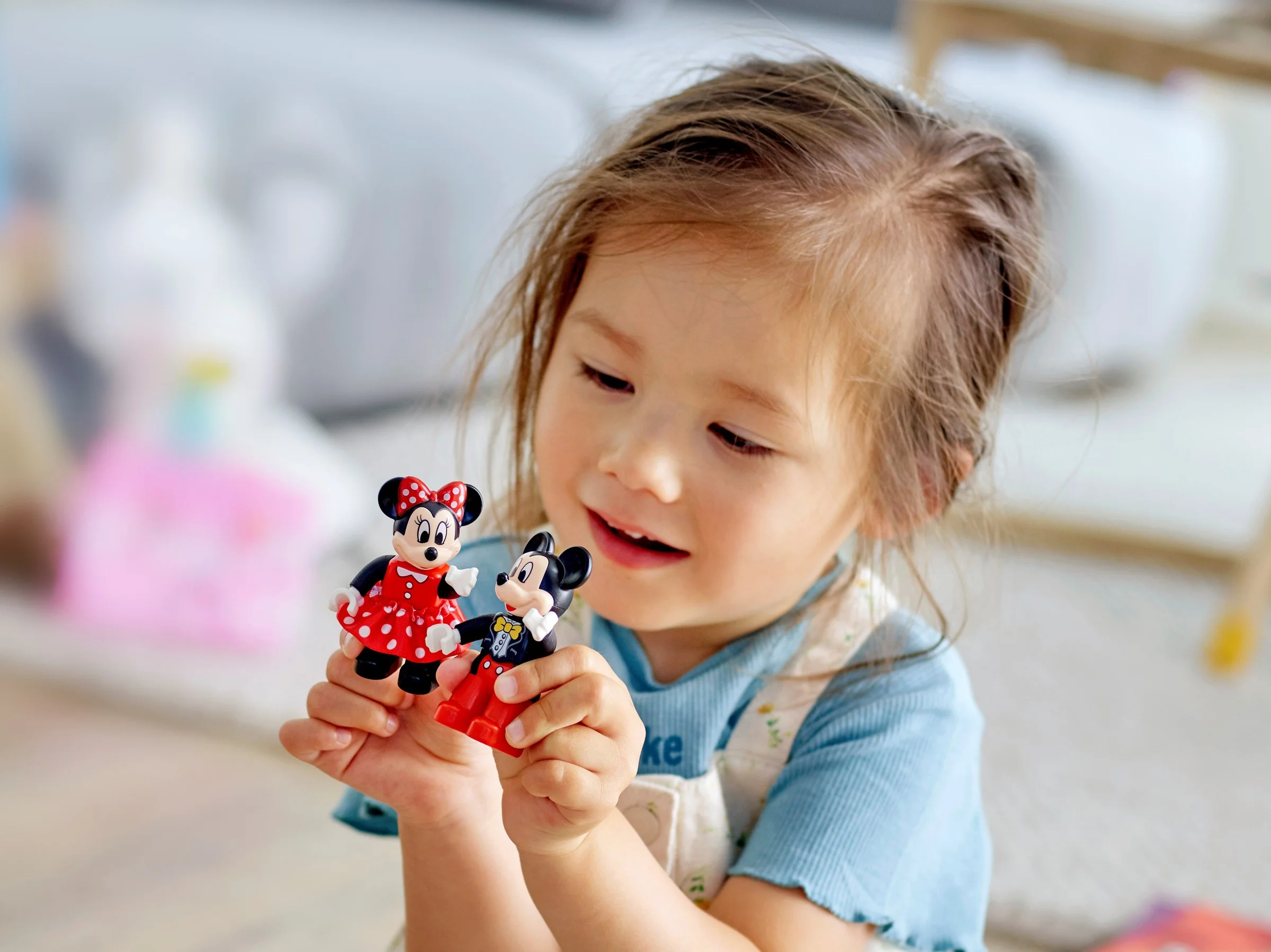 Fetiță zâmbitoare ținând figurinele Mickey și Minnie, jucându-se cu trenulețul aniversar Duplo.