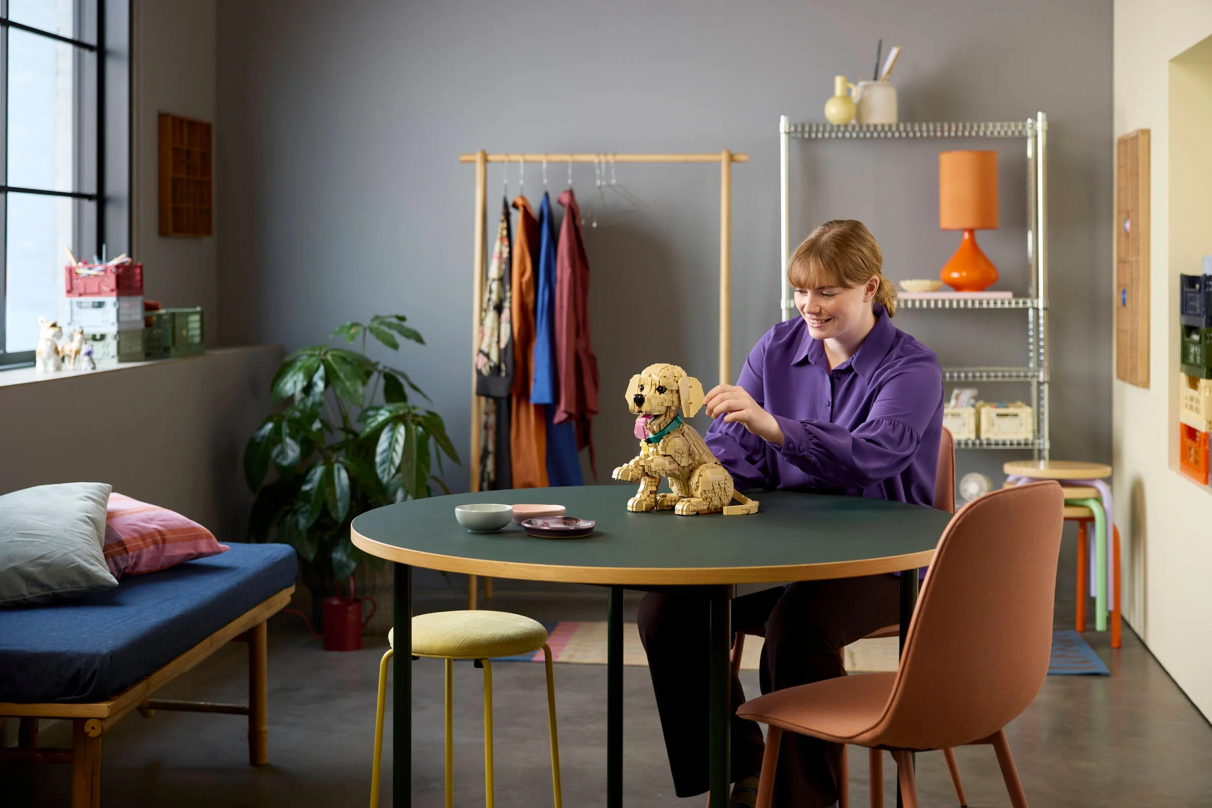 O femeie interacționează cu modelul LEGO Golden Retriever Puppy pe o masă, într-o cameră luminoasă.