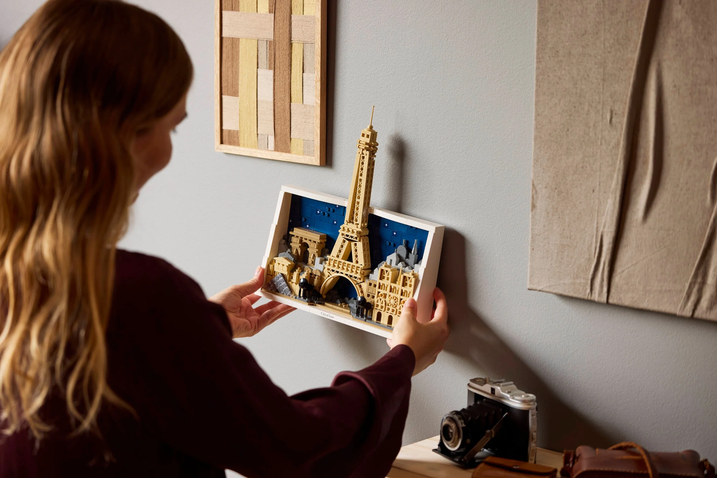 O femeie aranjează cadrul LEGO cu Parisul pe perete. Cadru alb, detaliat, turn Eiffel înalt.