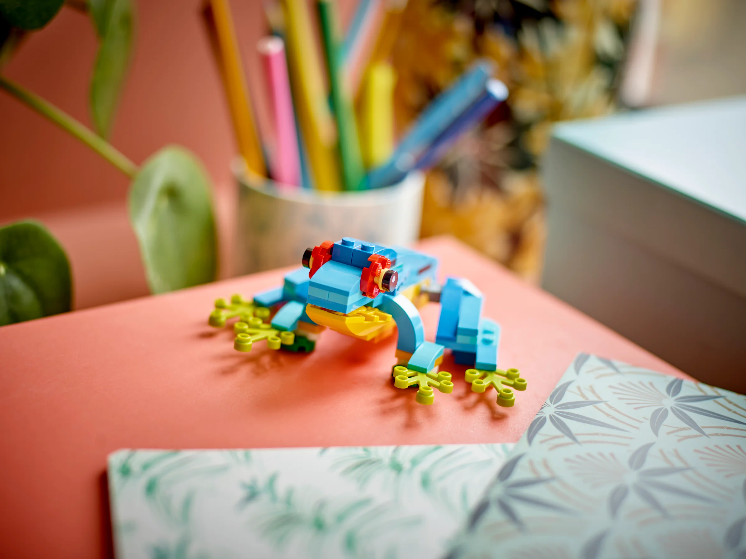 O broască colorată LEGO stă pe o suprafață portocalie, într-un cadru artistic și viu.