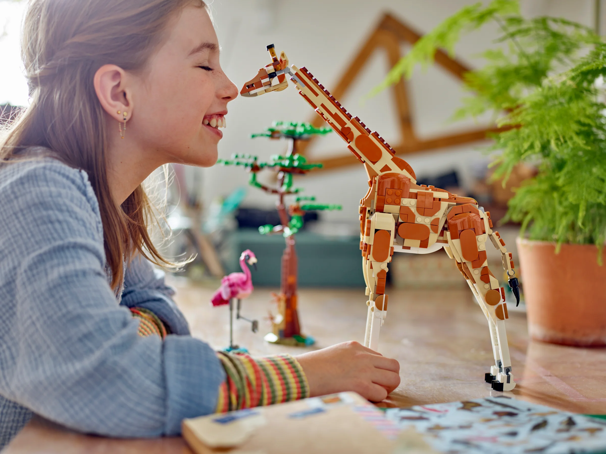 O fată zâmbește atingând girafa LEGO. Setul include un flamingo și un copac din cărămizi.