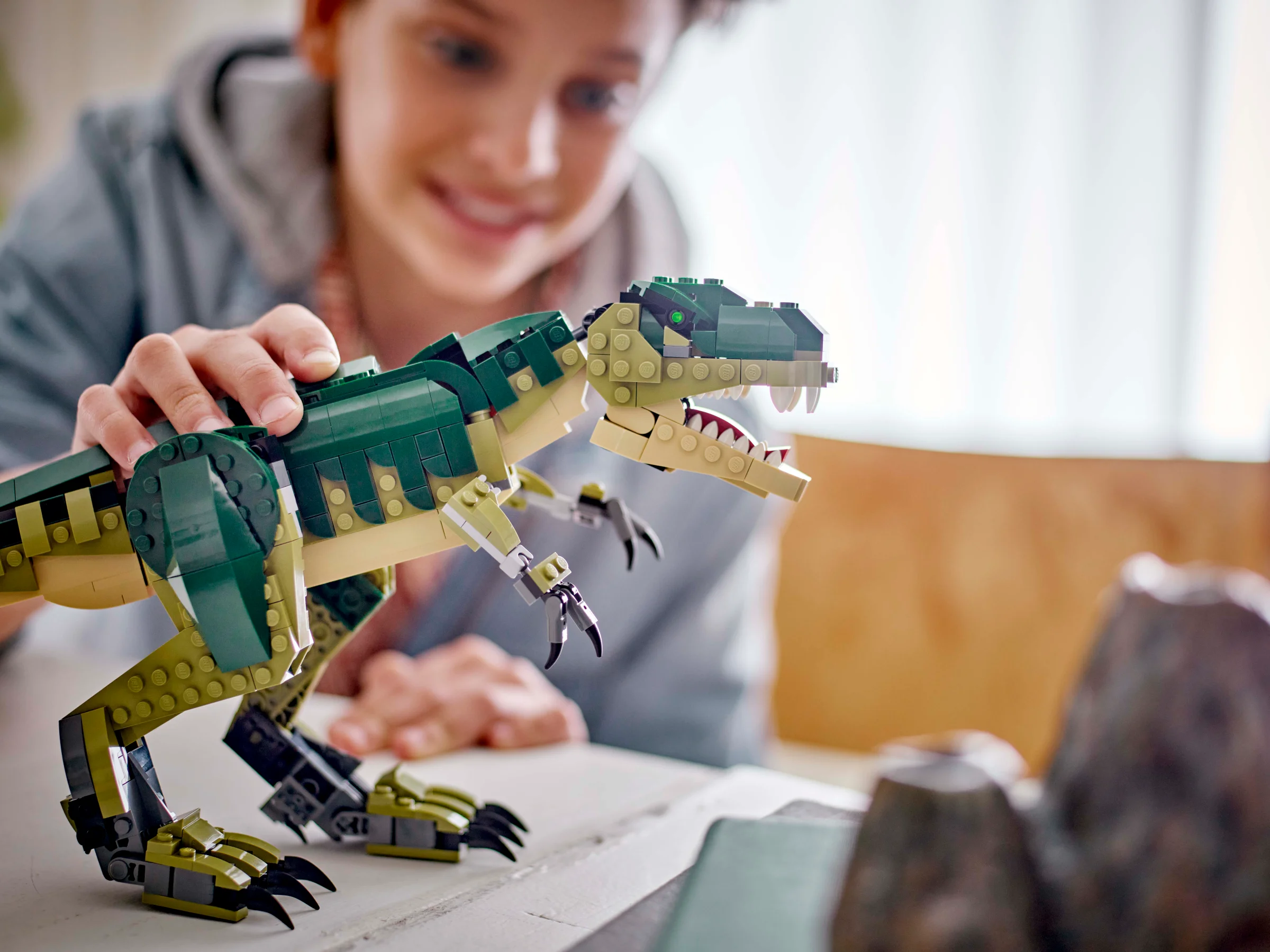 Un băiat zâmbitor ține un T. rex LEGO verde și bej, privind modelul cu atenție.