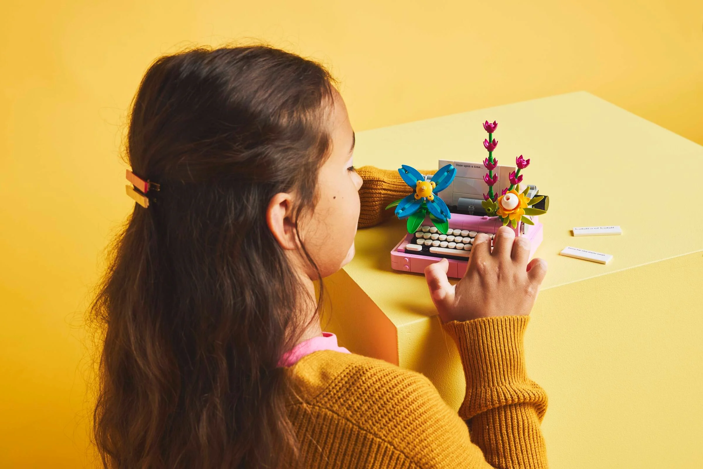 Fată interacționând cu mașina de scris LEGO, cu flori decorative. Fundal galben.