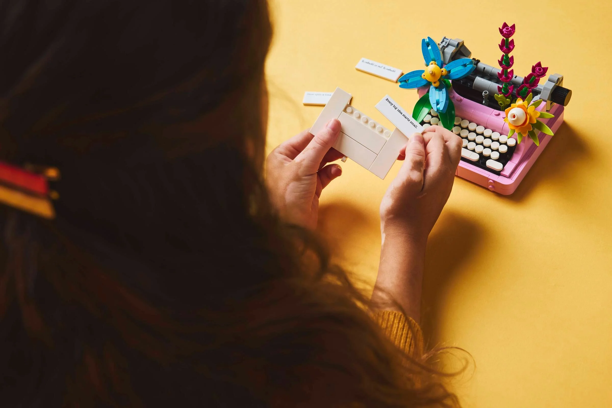 Modelul LEGO al unei mașini de scris roz, cu flori, și o persoană interacționând cu mesajul scris.
