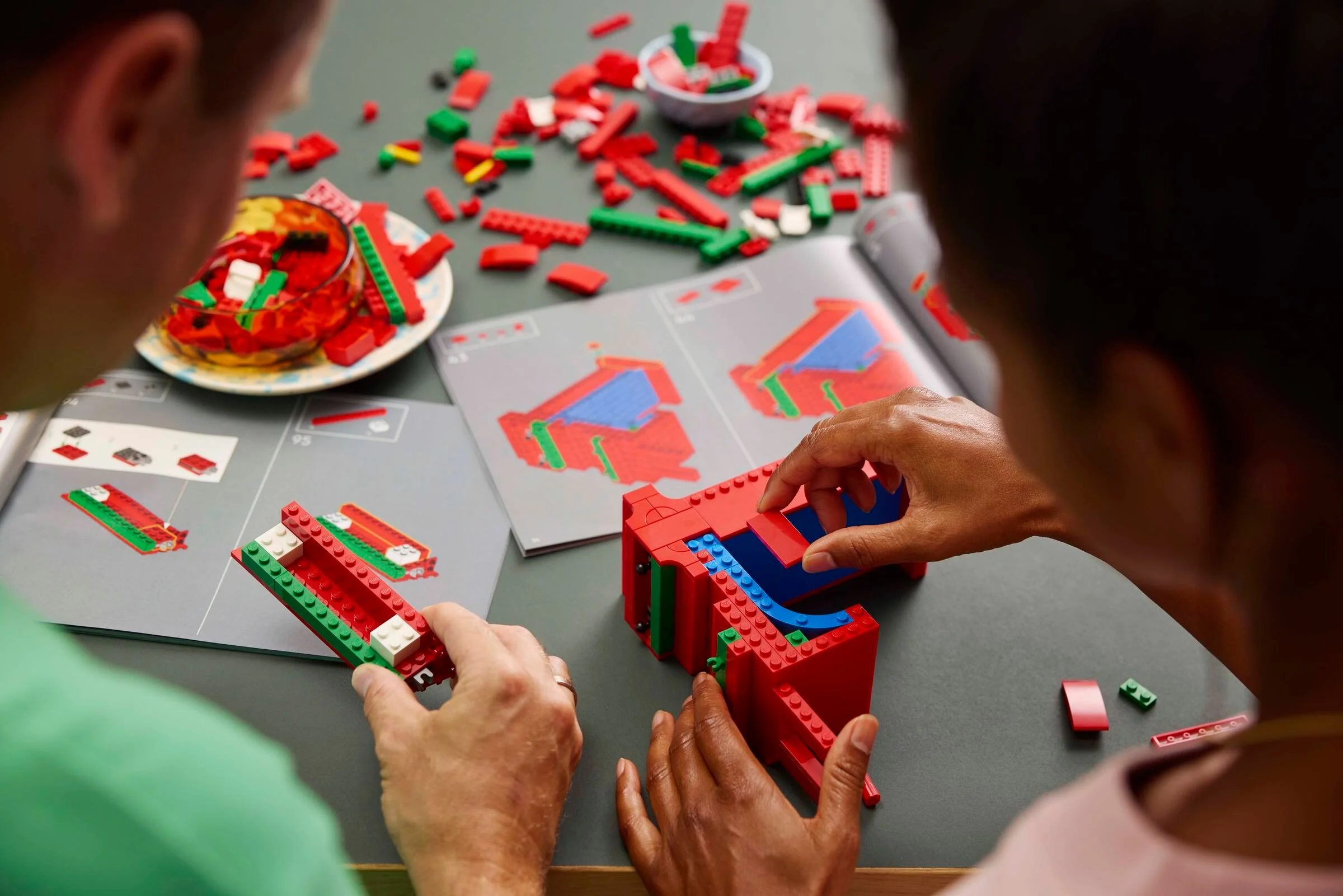 Două persoane construiesc setul LEGO Love, urmând instrucțiunile. Piese roșii și verzi împrăștiate.