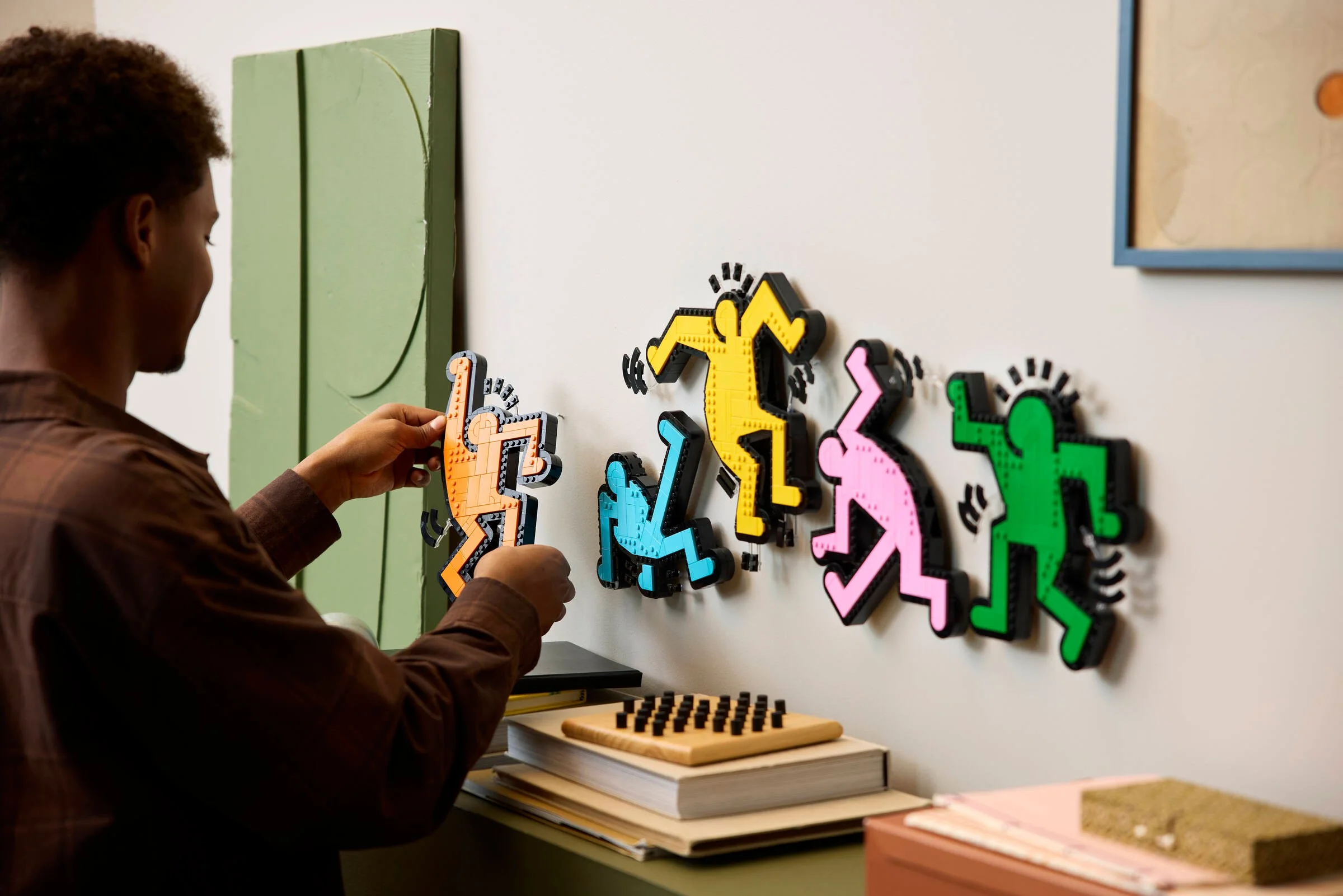 Imagine cu o persoană montând figurinele LEGO Keith Haring pe un perete alb.