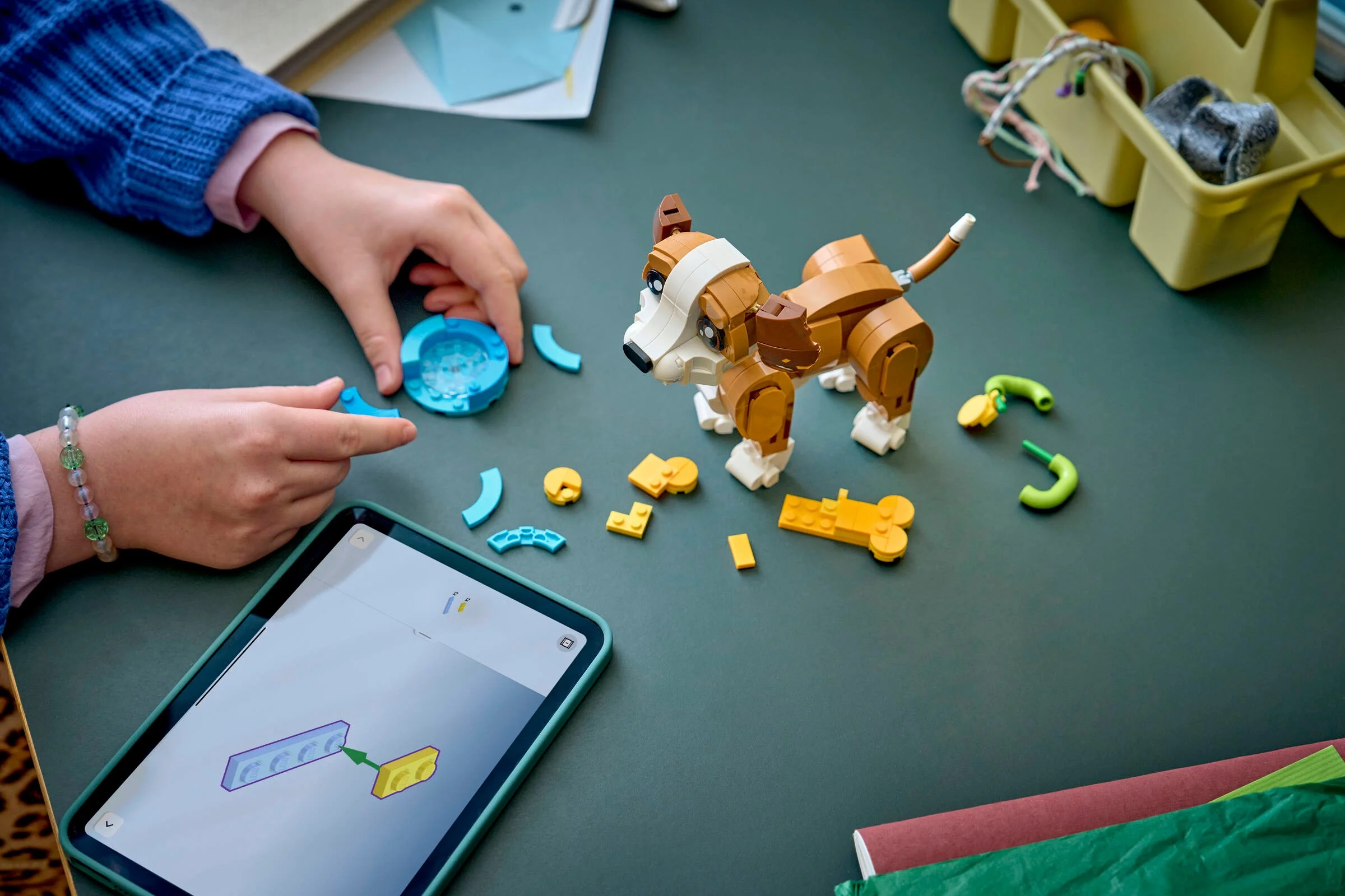 Copil construind cățelul LEGO. Instrucțiuni pe tabletă. Piesele sunt împrăștiate pe masă.