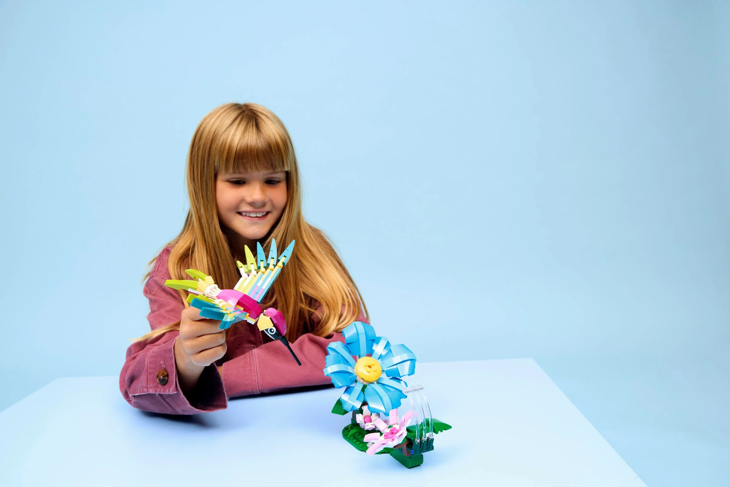 Fetiță zâmbind cu colibri LEGO colorat și floare, pe un fundal albastru pastel.