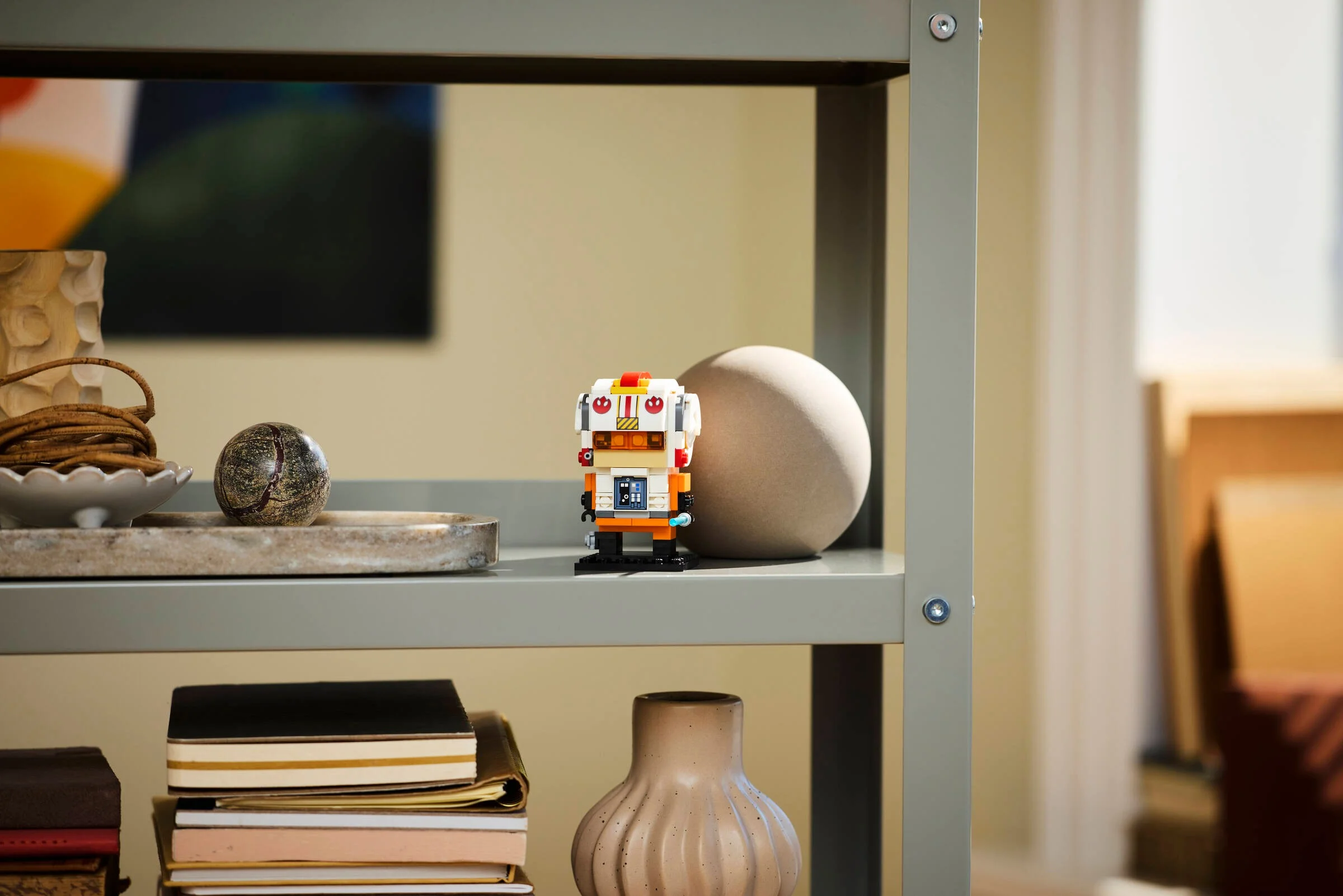 O figurină BrickHeadz Luke Skywalker pilot rebel, așezată pe o etajeră, decorând camera.