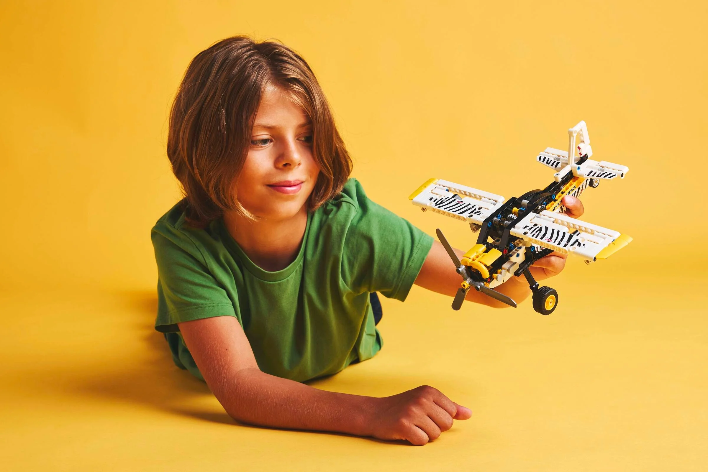 Un copil se joacă cu avionul LEGO Technic. Fundal galben simplu.