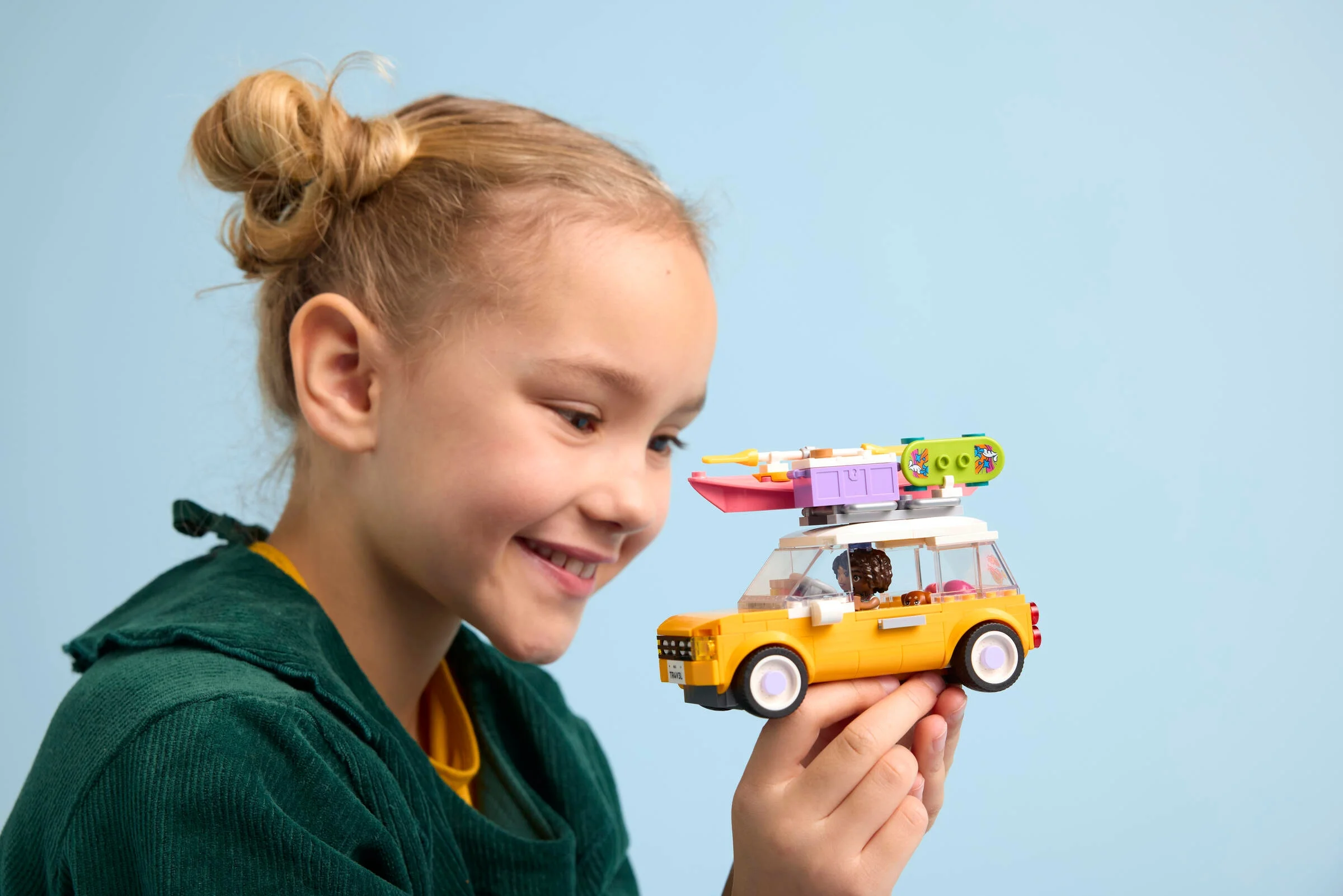 Fetiță zâmbitoare admirând mașina de călătorie LEGO Friends, galbenă și plină de accesorii.