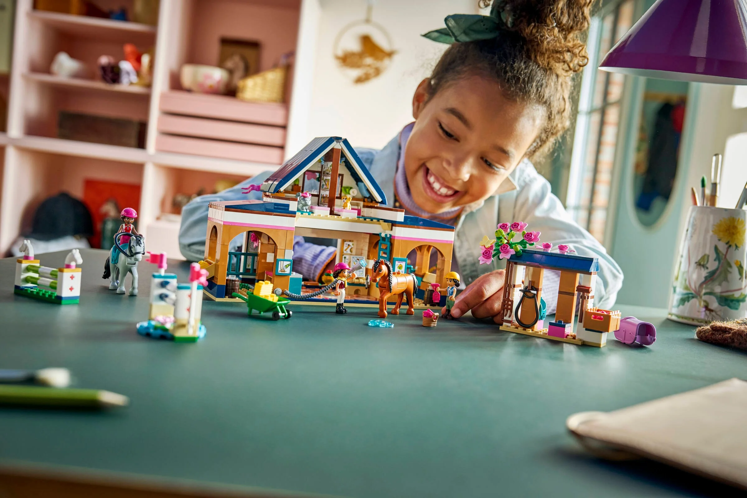 O fetiță zâmbește jucându-se cu setul LEGO Friends 42688 Academie de Echitație. Detalii și minifigurine vizibile.