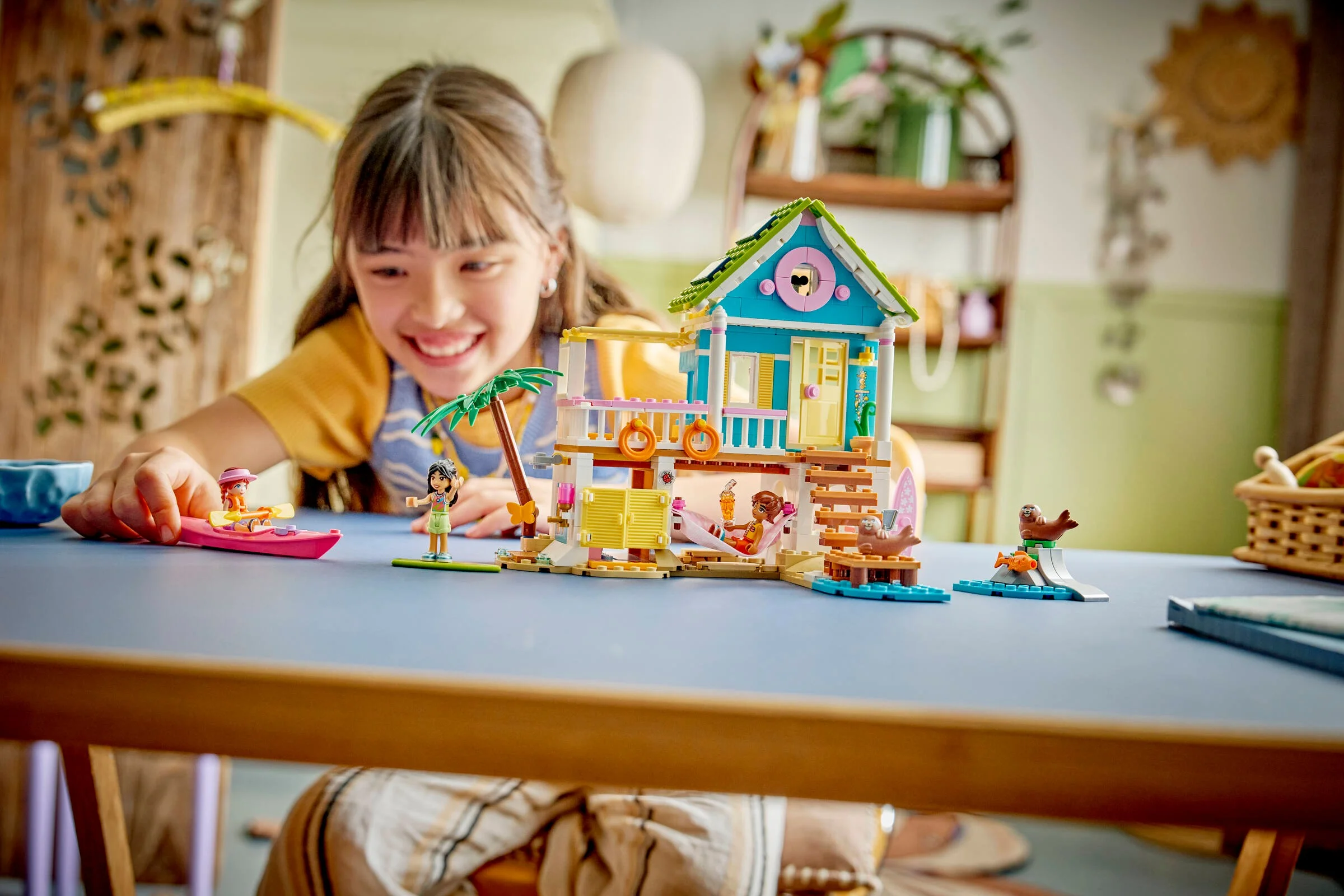 Fetiță zâmbitoare se joacă cu setul LEGO 42699, căsuța de plajă cu foci, pe o masă albastră.