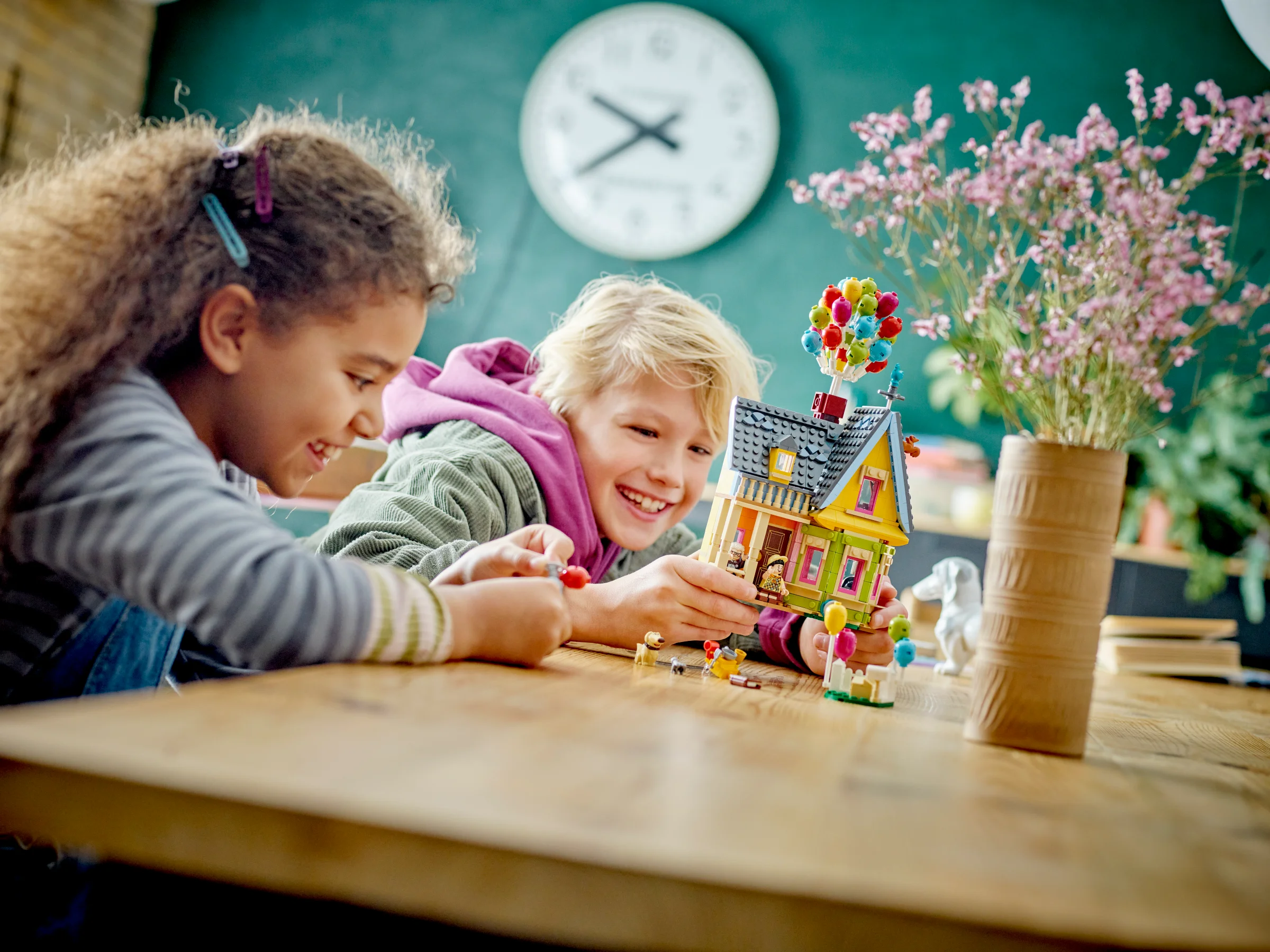 Copii zâmbind jucându-se cu casa 'Up' LEGO 43217 pe o masă de lemn.