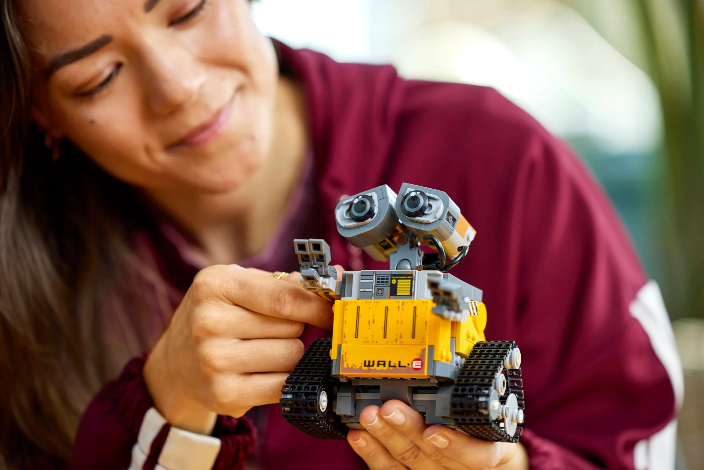 Femeie zâmbind ținând setul LEGO WALL-E și EVE. Modelul este focalizat, fundalul ușor neclar.