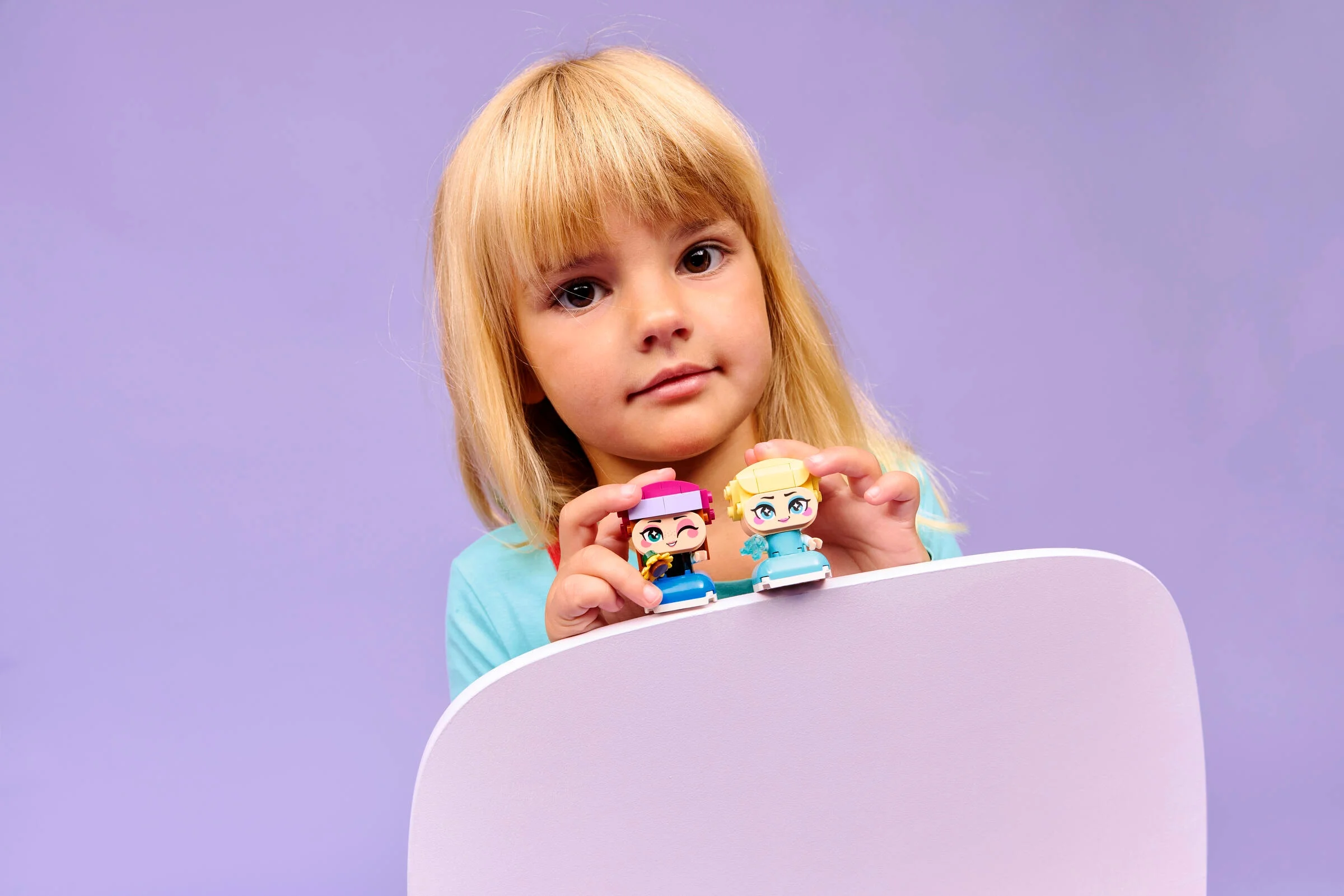 Copil ținând minifigurinele Anna și Elsa, zâmbind. Fundal mov.