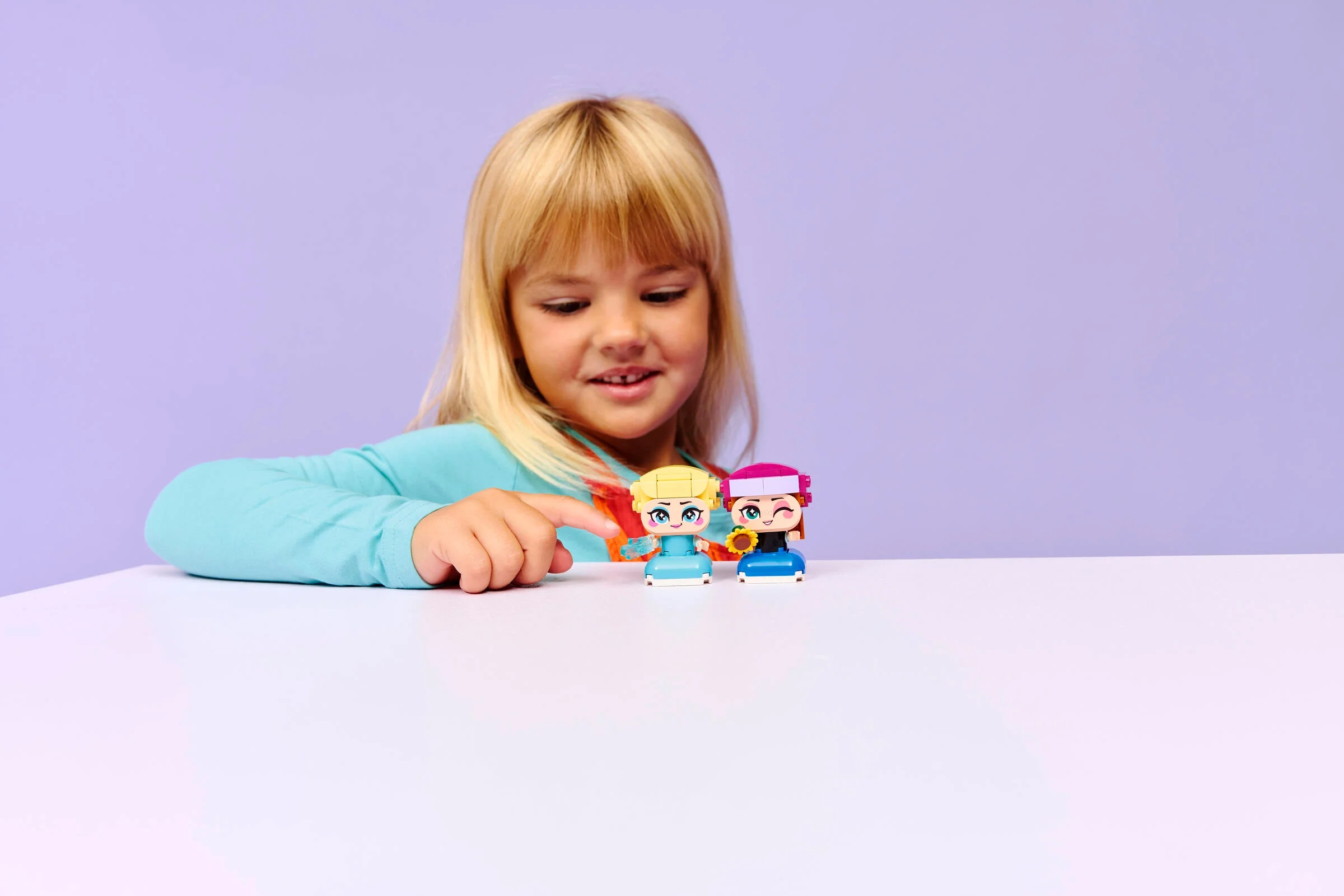 Fetiță interacționând cu figurinele LEGO Anna și Elsa, pe o masă albă.
