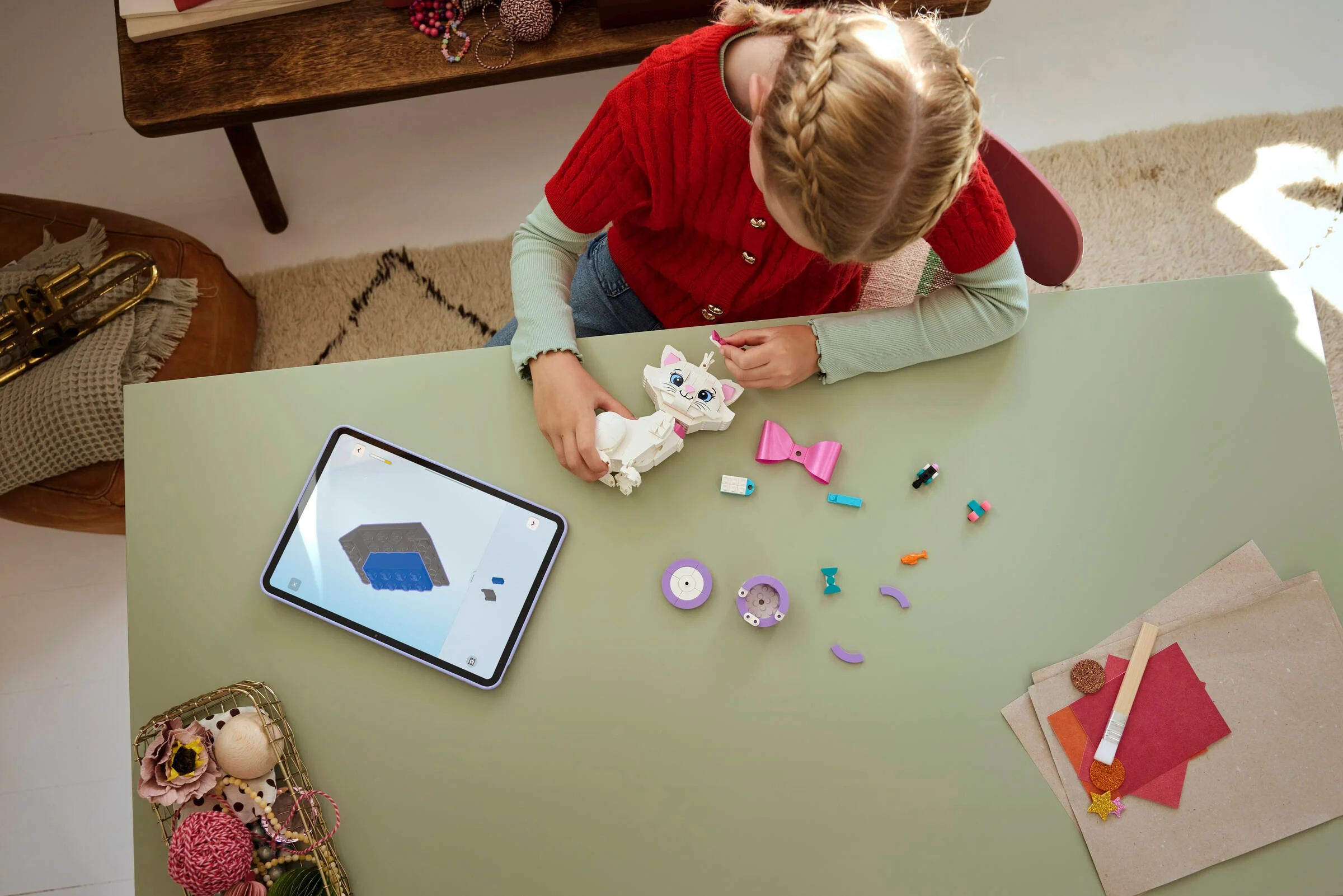 O fetiță construiește LEGO Marie urmând instrucțiunile de pe tabletă. Piesele sunt aranjate pe masă.
