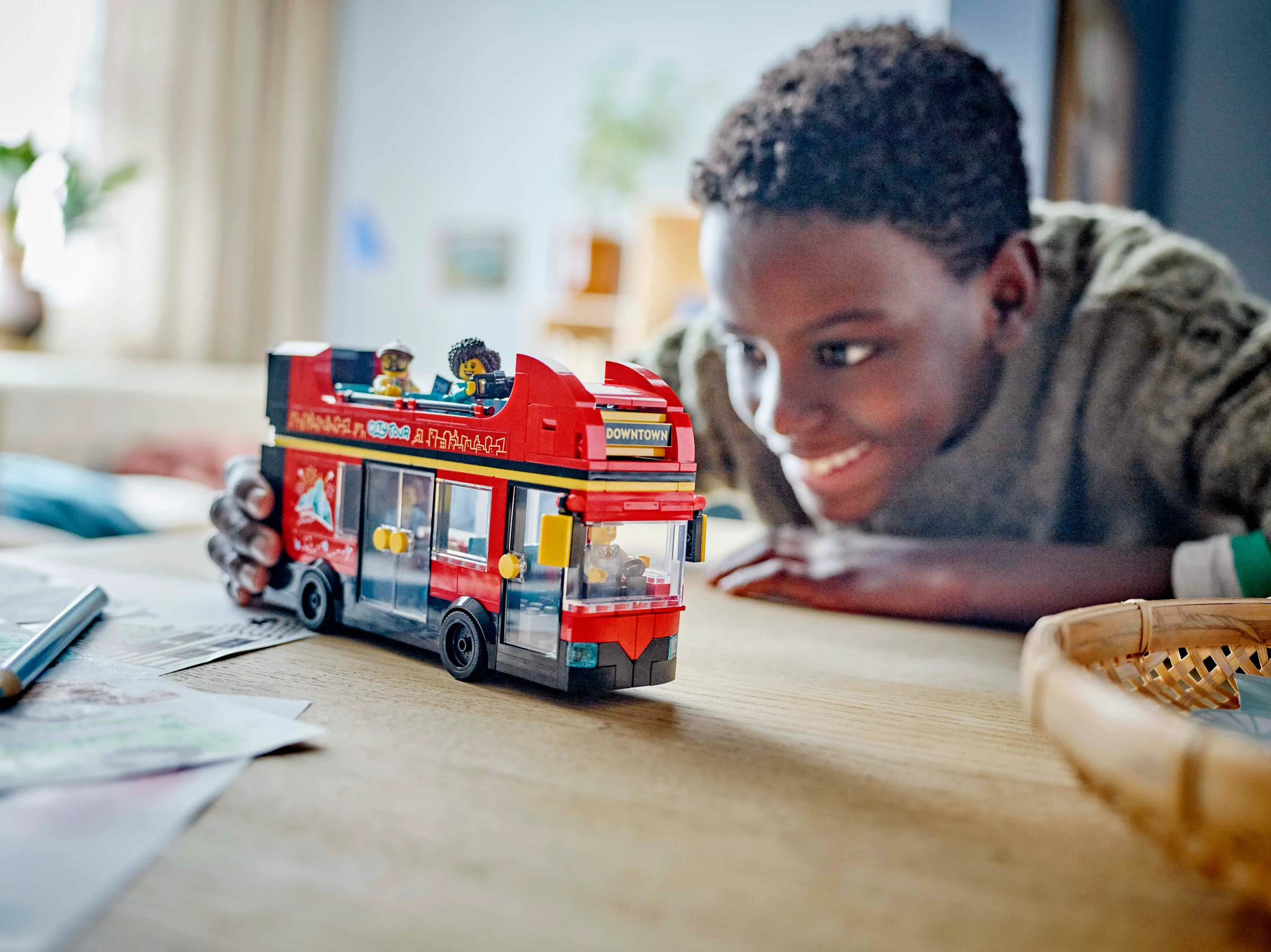 Băiat zâmbind admirând autobuzul turistic roșu LEGO City, cu minifigurine la bord.
