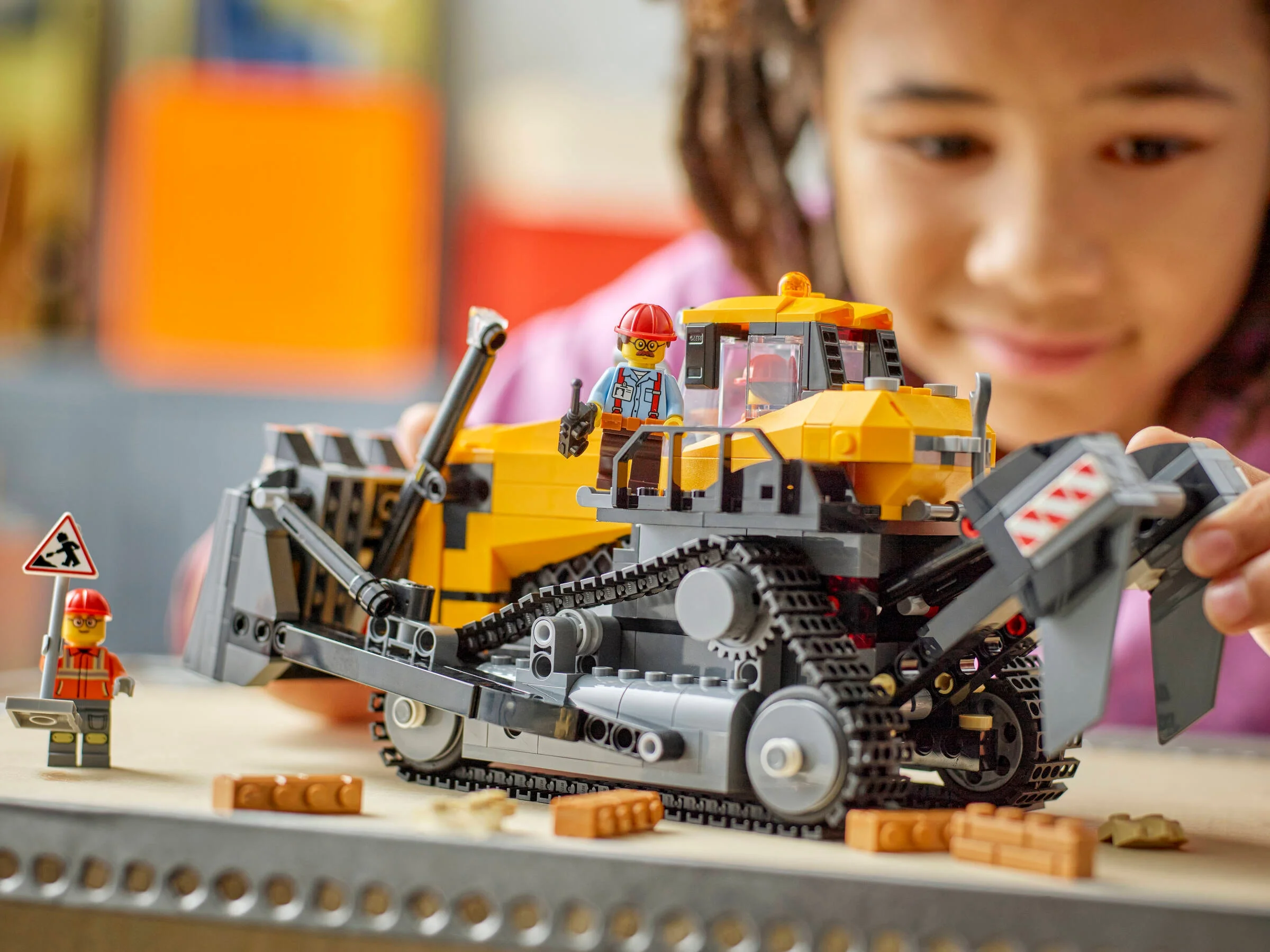 Buldozerul galben și gri LEGO City este prezentat, cu detalii și o fetiță în fundal.