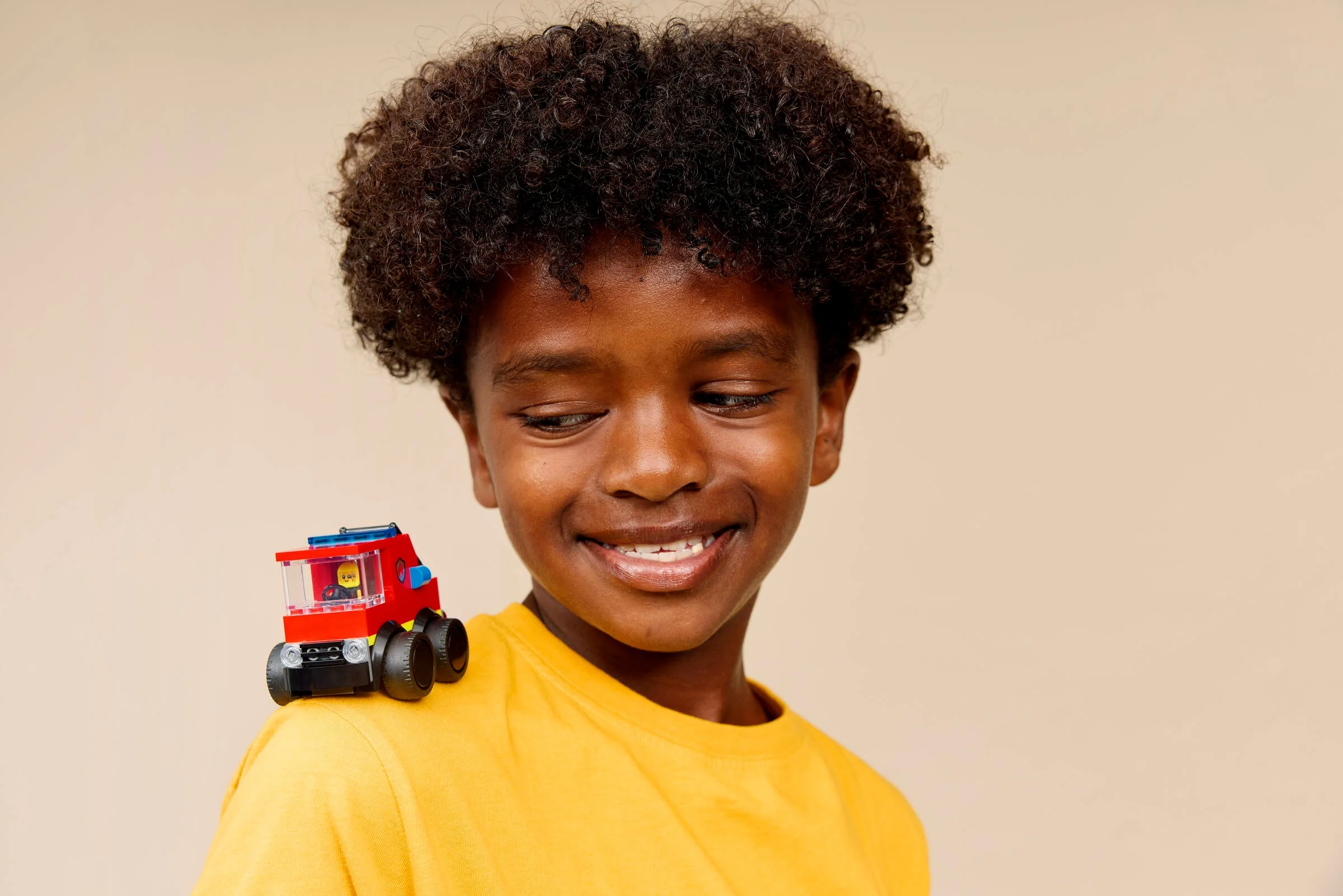 Un băiat zâmbitor cu o mașină de pompieri LEGO pe umăr.