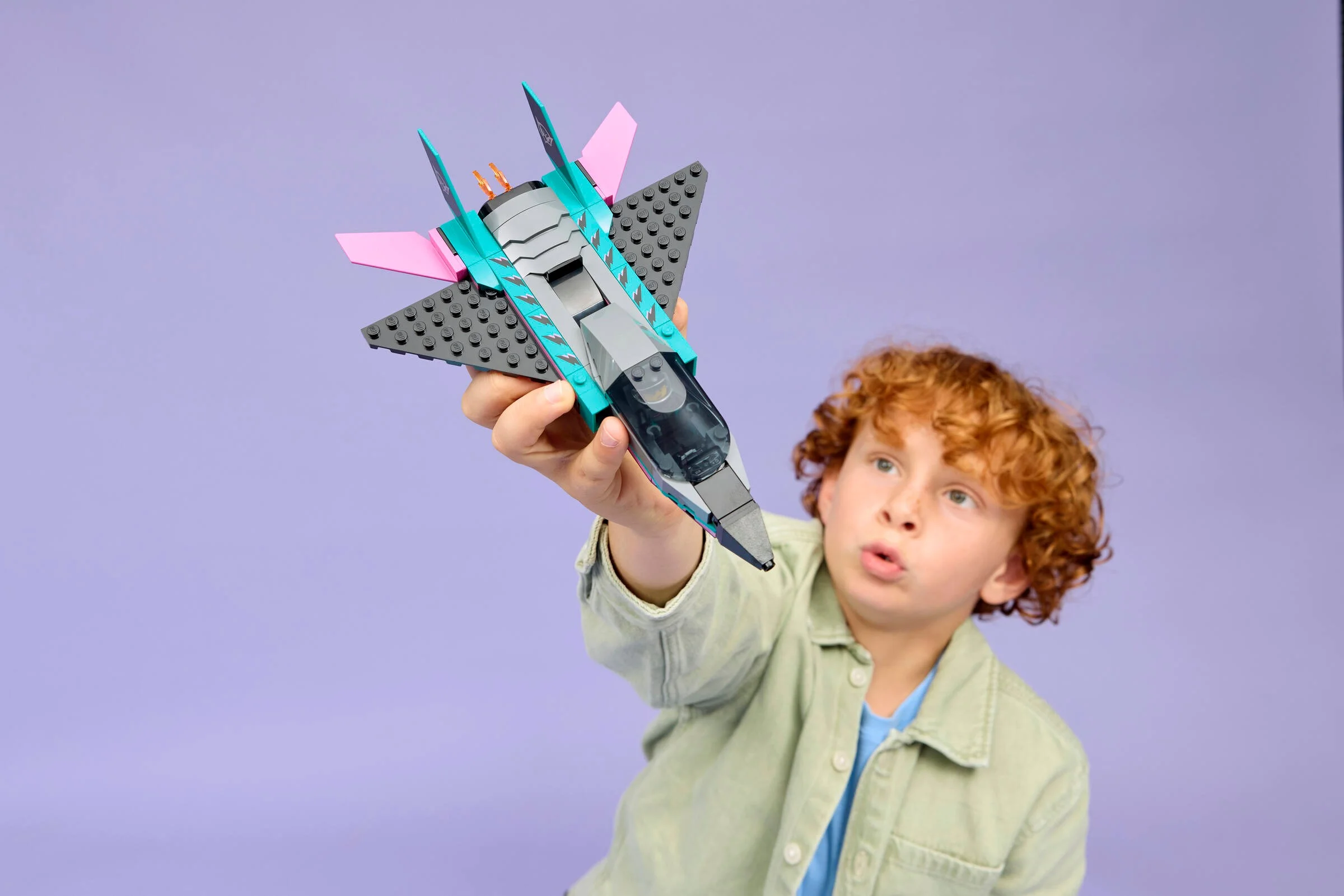 Un copil ține avionul LEGO 60489, cu detalii roz și turcoaz, pe fundal mov.