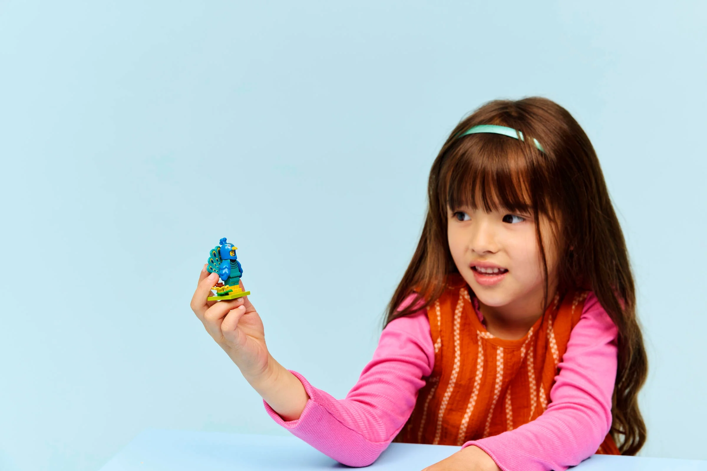 O fetiță admiră minifigurina Costum de Păun LEGO 71051, pe fundal albastru deschis.