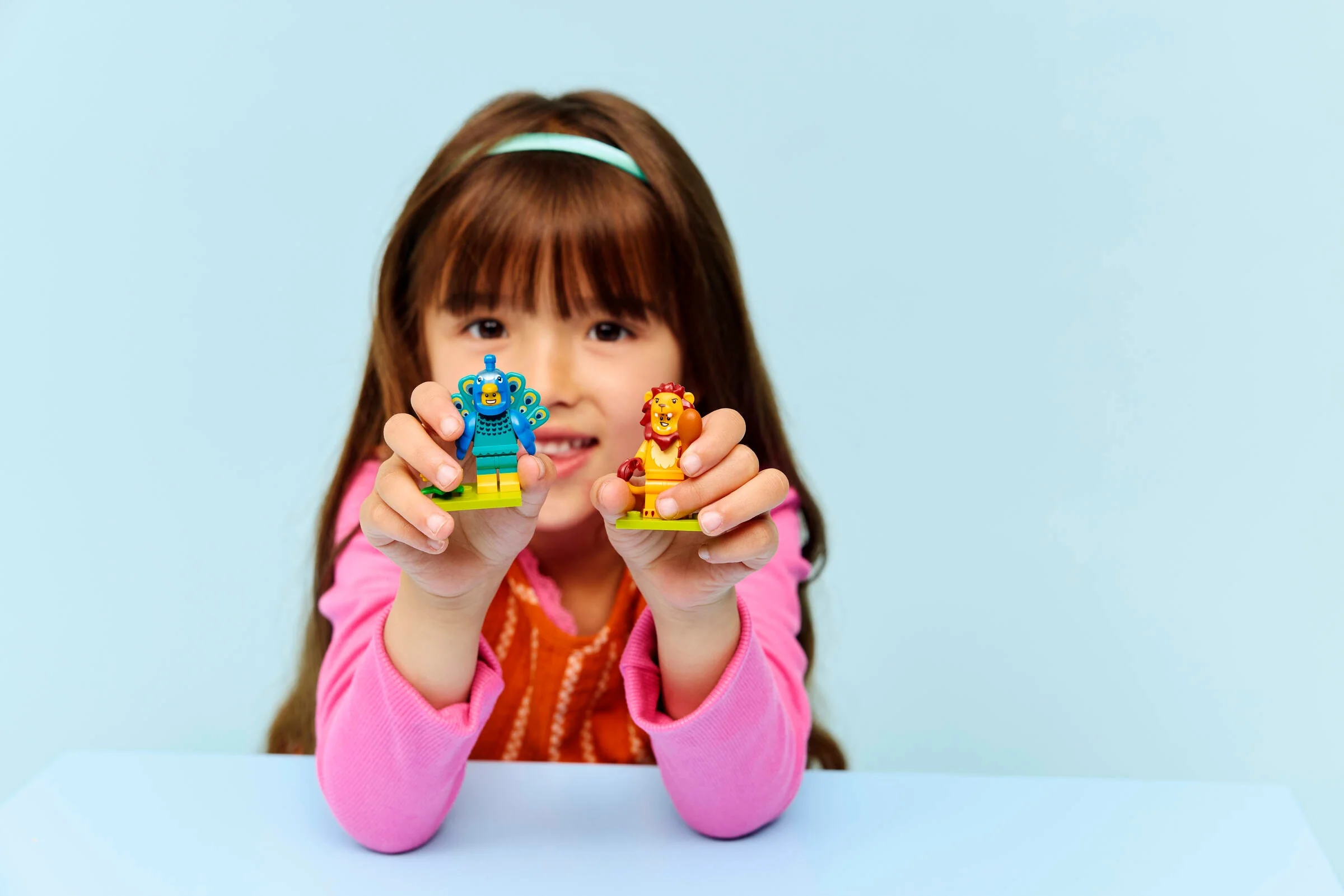 O fetiță prezintă minifigurinele LEGO Păun și Leu, zâmbind. Fundal albastru.