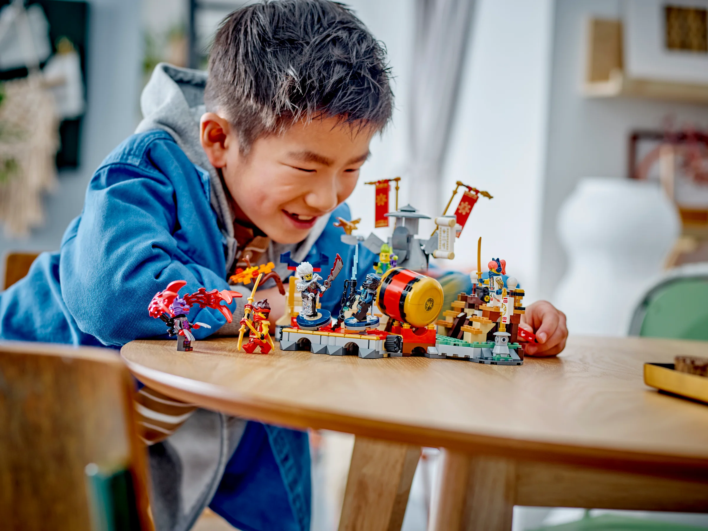 Copil zâmbind, jucând cu setul LEGO Ninjago Arena de Turneu 71818 pe o masă.
