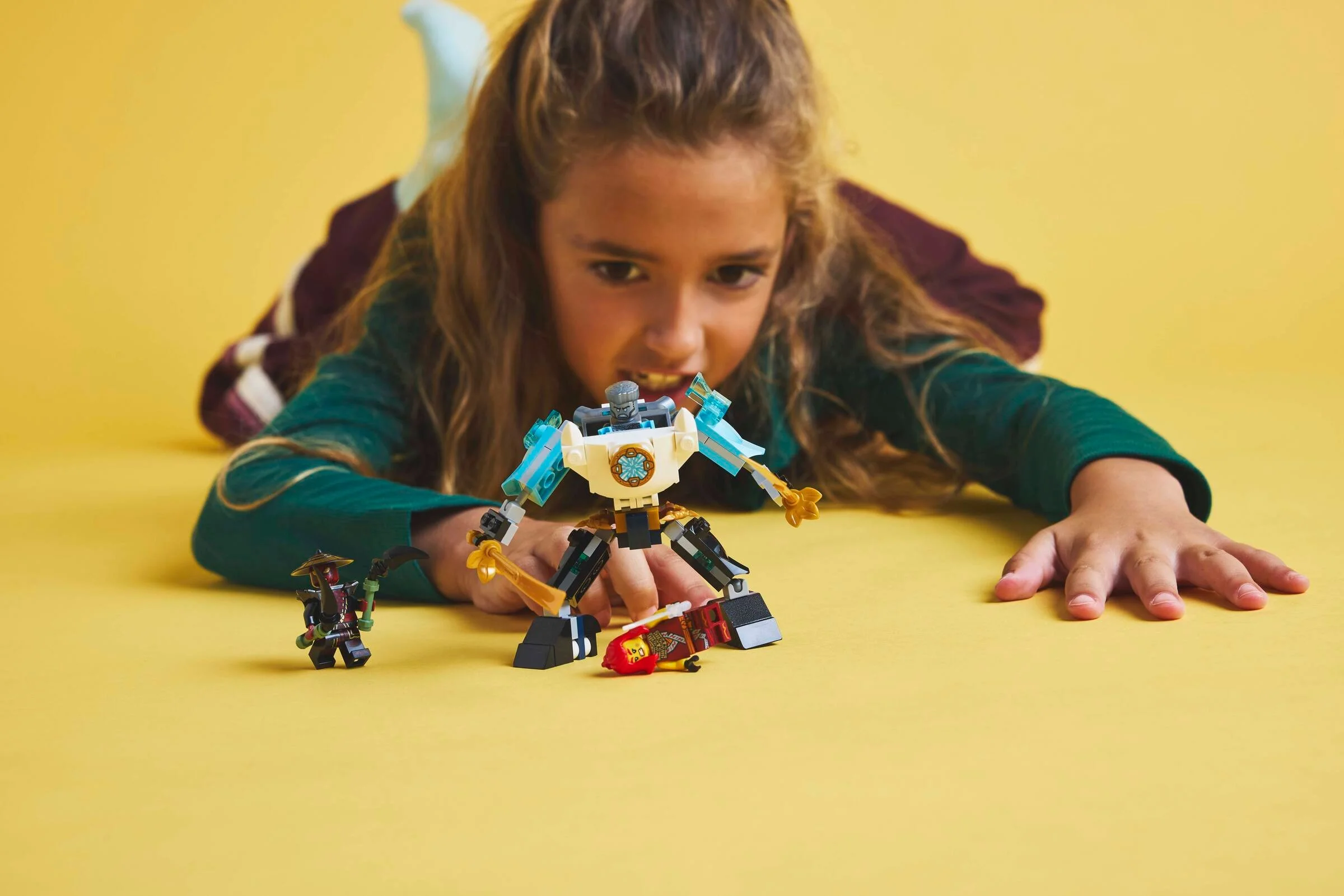 Copil se joacă cu Zane's Battle Suit Mech, personajele și placa Ninjago pe un fundal galben.