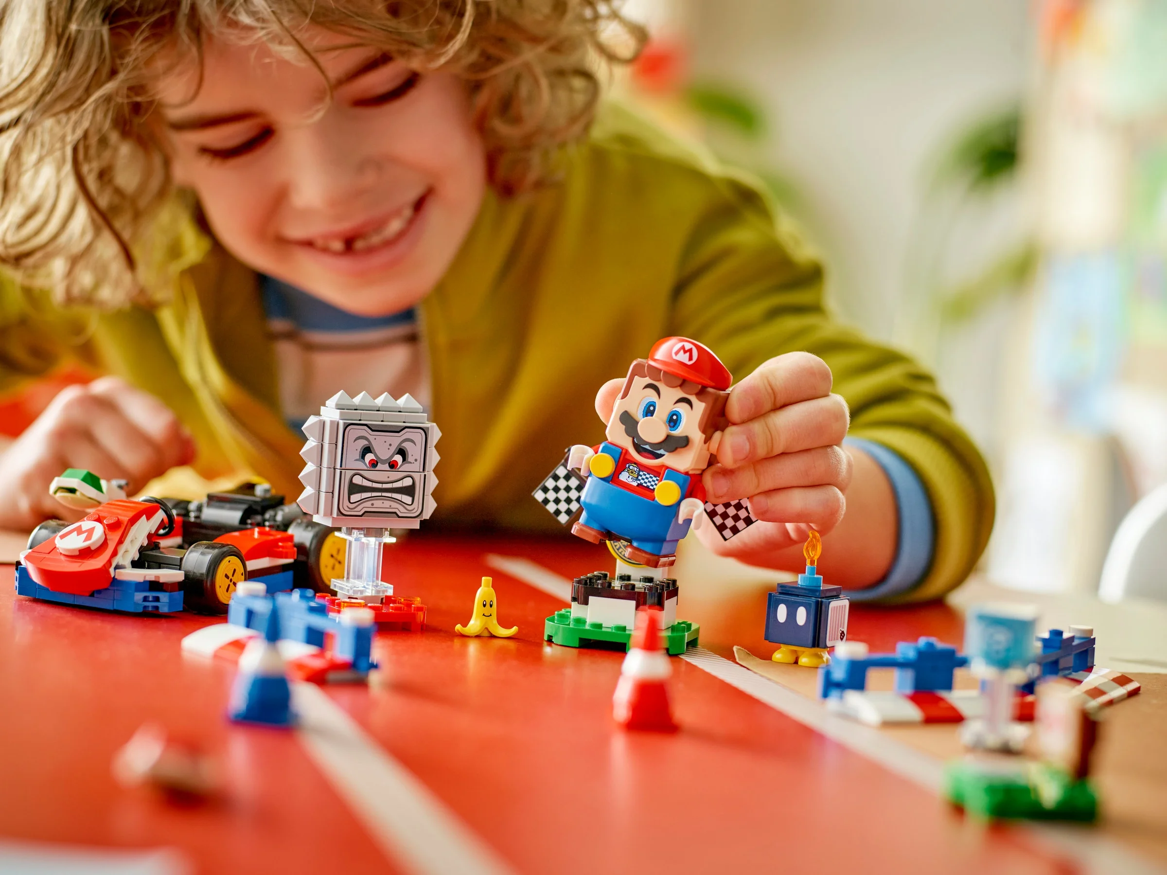 Copil zâmbitor se joacă cu setul LEGO Mario Kart pe o masă roșie.