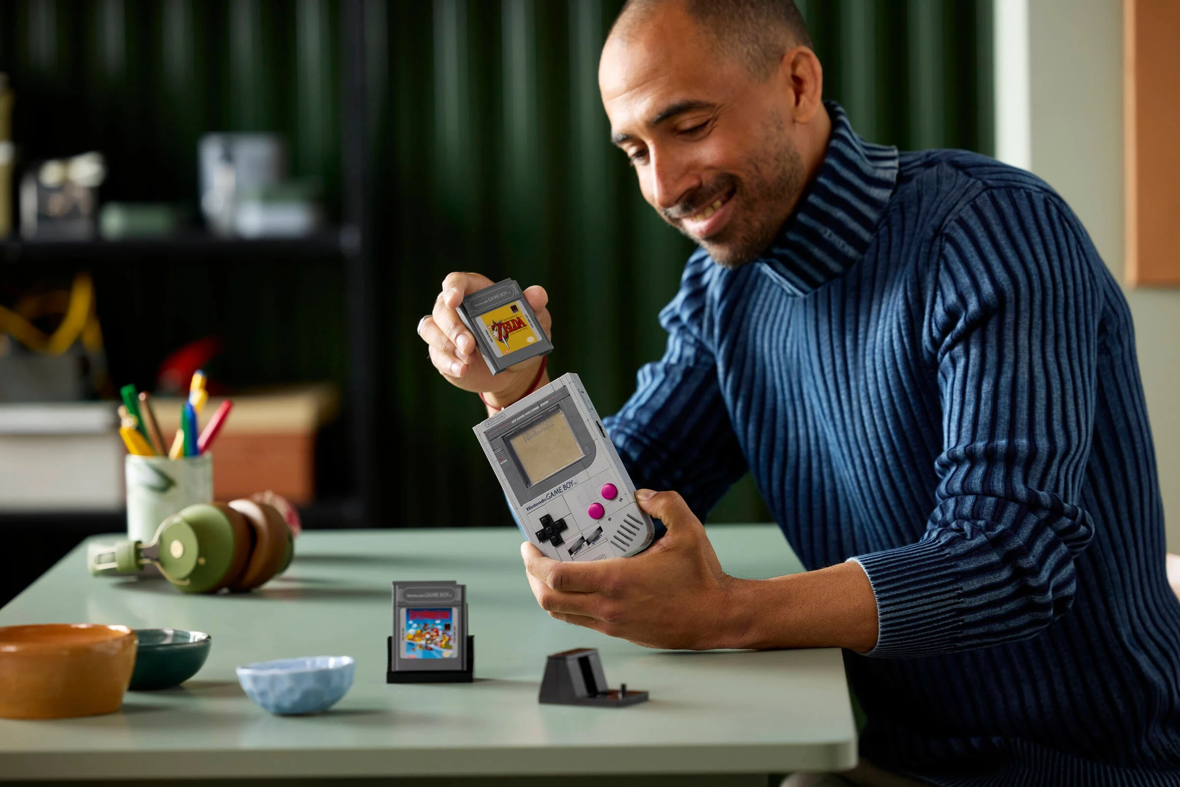 Un bărbat zâmbind admiră Game Boy-ul LEGO și cartușul jocului Zelda.