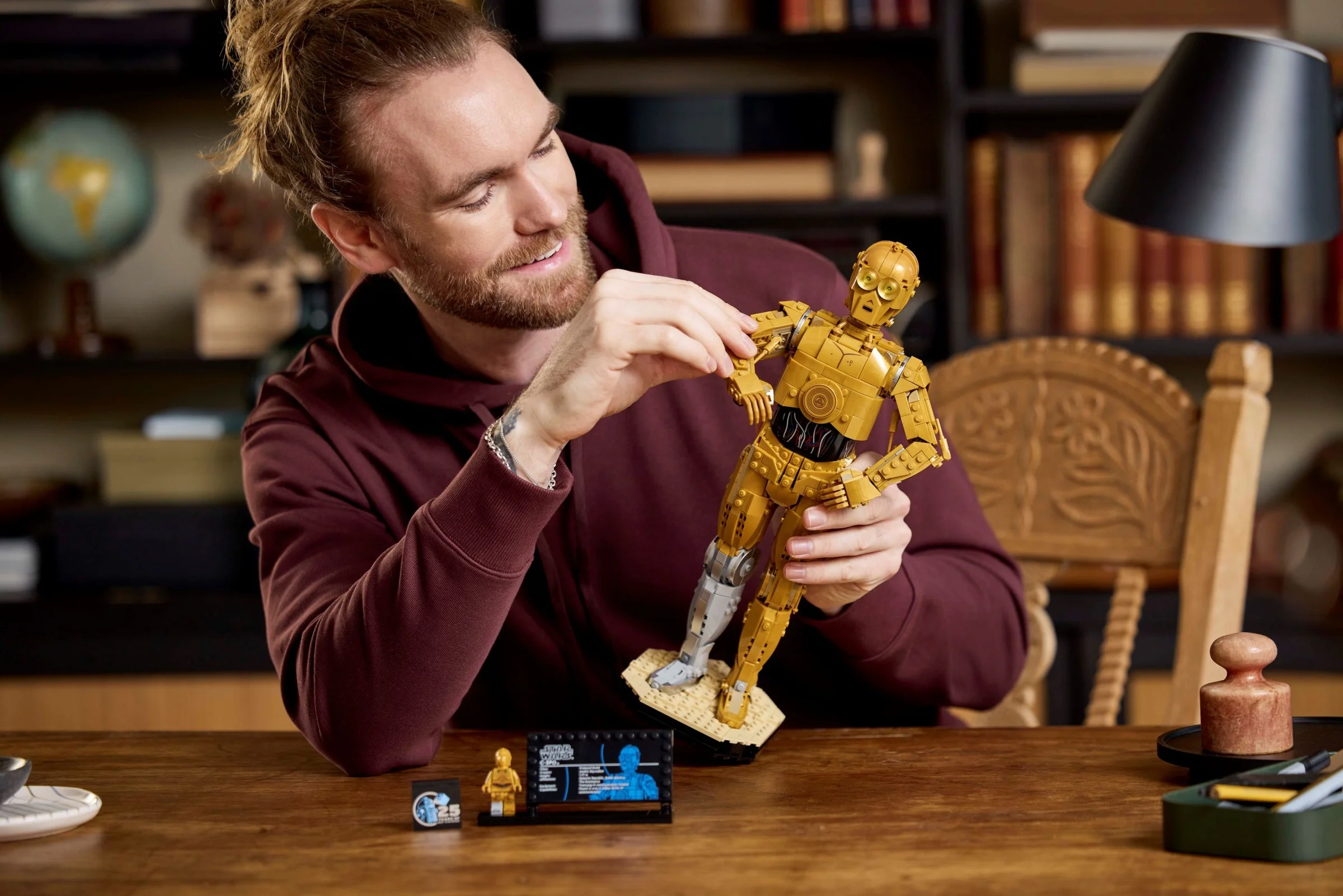 Un bărbat interacționează cu figurina C-3PO LEGO, analizând detaliile. Cadru cald și primitor.